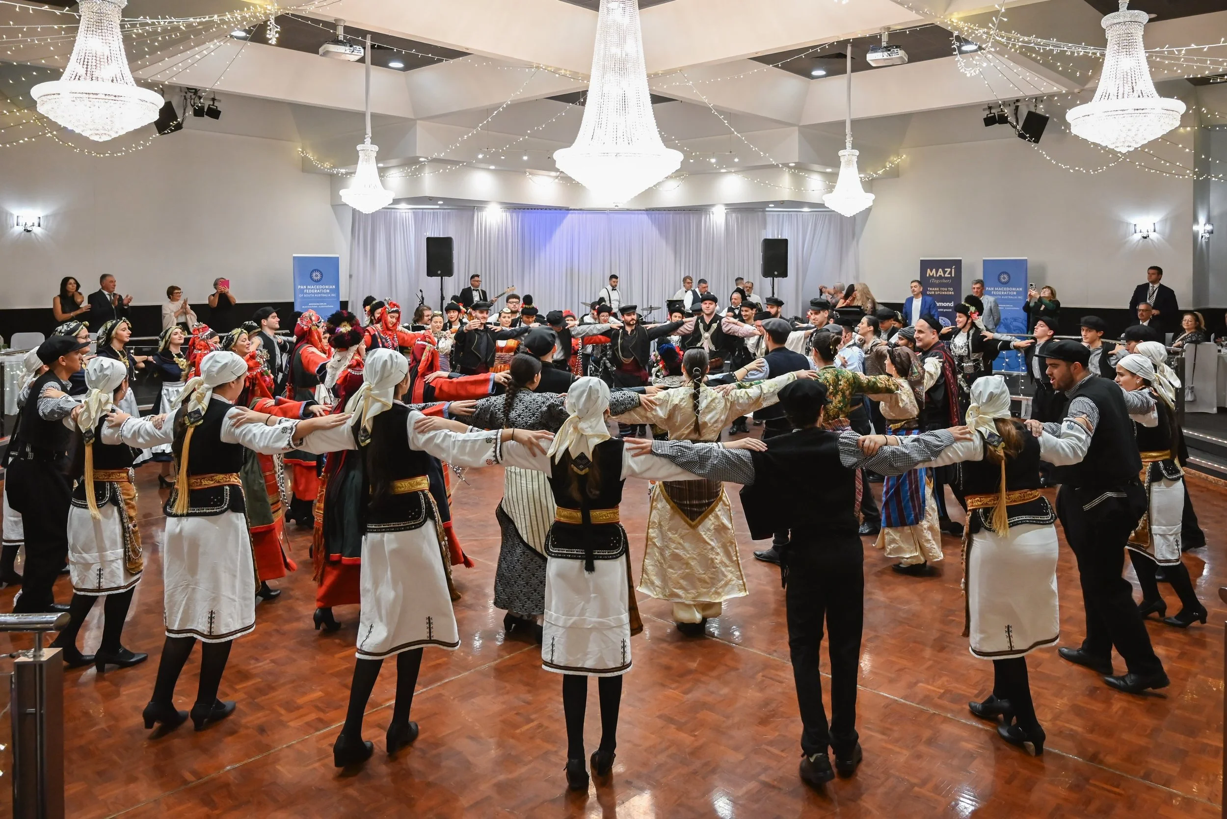 A large group of people in traditional Balkan attire dance in a circle during an indoor cultural event, with some onlookers clapping and taking photos. The room is decorated with Elegant chandeliers and string lights, and banners on the sides.