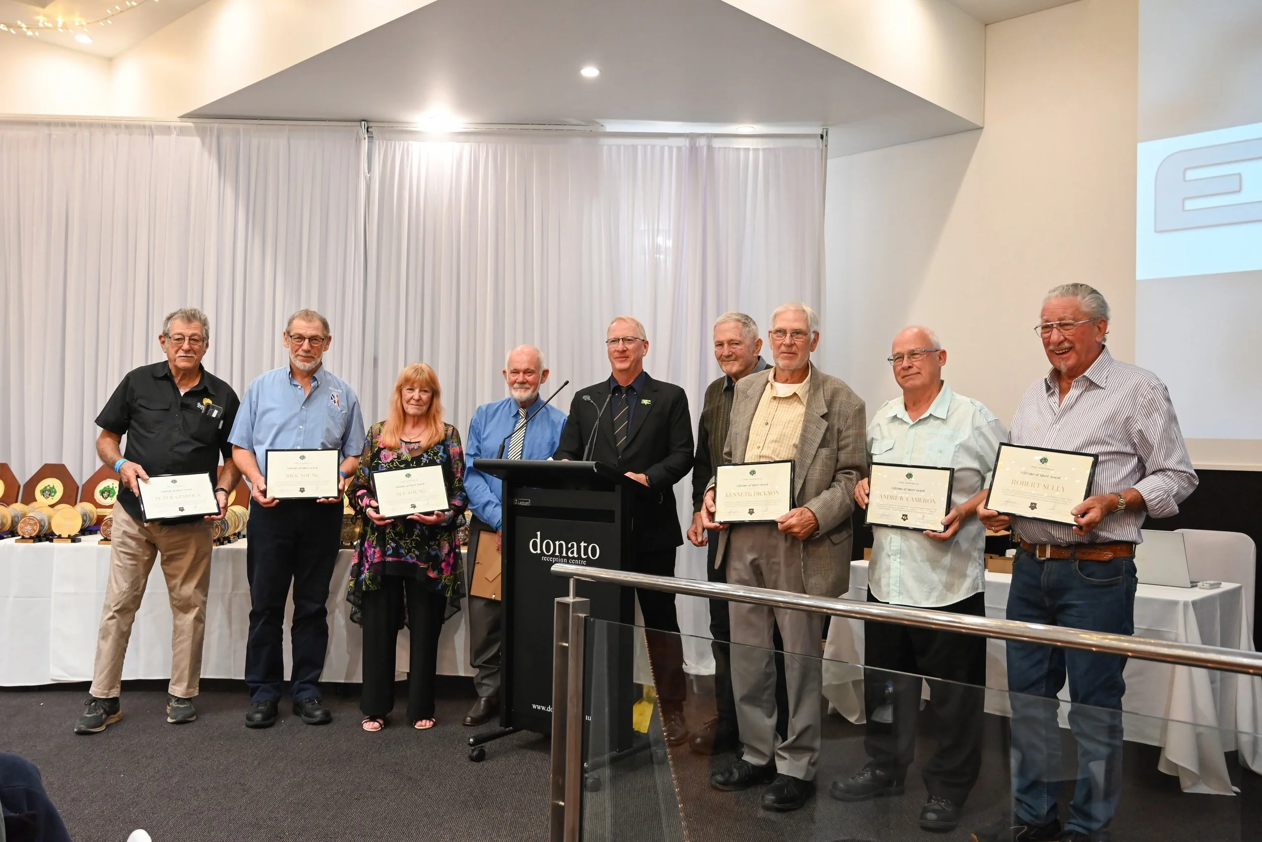A group of older adults standing in a line, holding certificates, at an award ceremony in a conference room with white curtains and a table with plaques and trophies in the background.