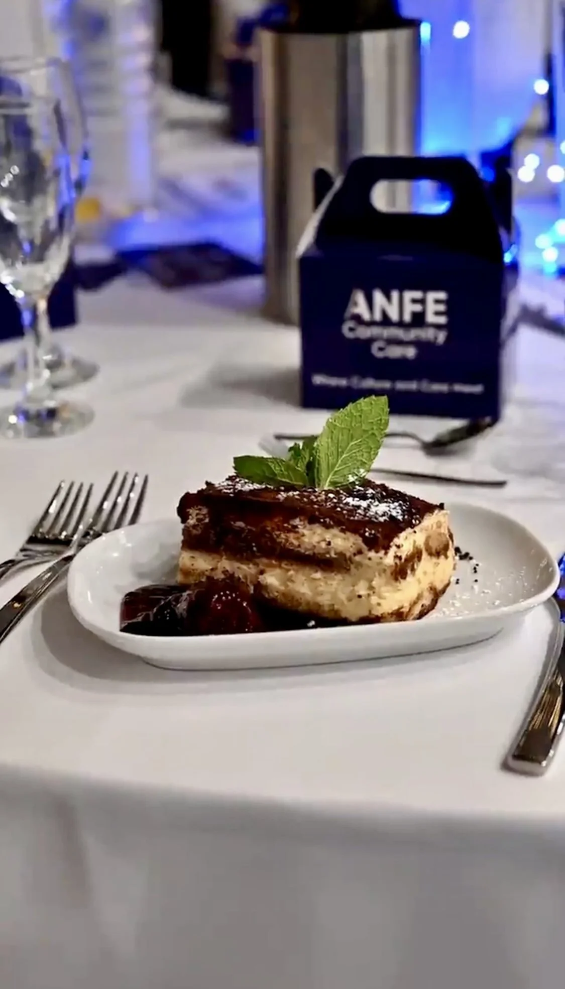 A slice of layered dessert topped with mint leaves on a white plate, set on a dining table with glasses, forks, and a box of ANFE Culinary Company Creme in the background.