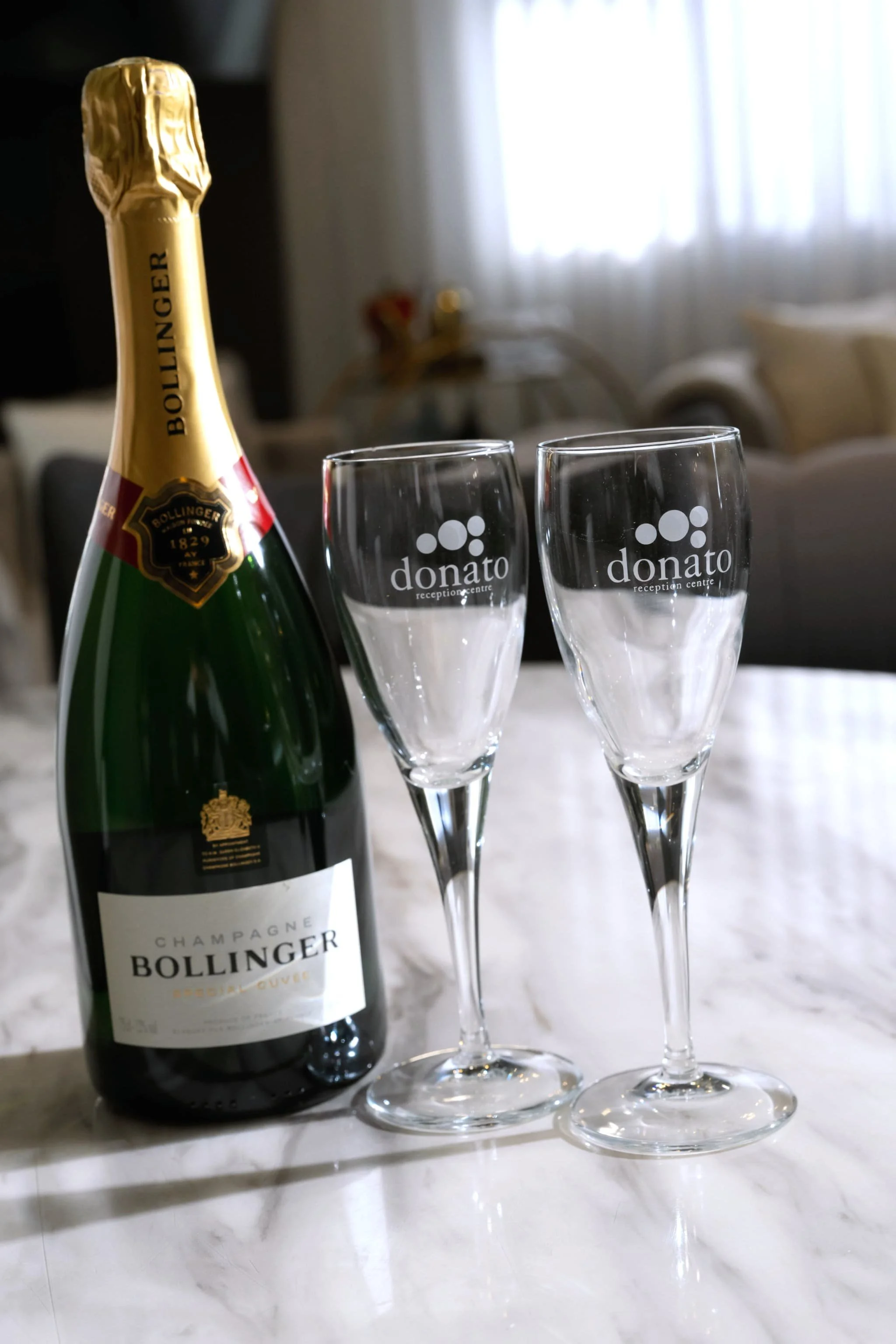 A bottle of Bollinger champagne and two empty champagne glasses on a marble surface, with a blurred indoor background including a sofa and room curtains.