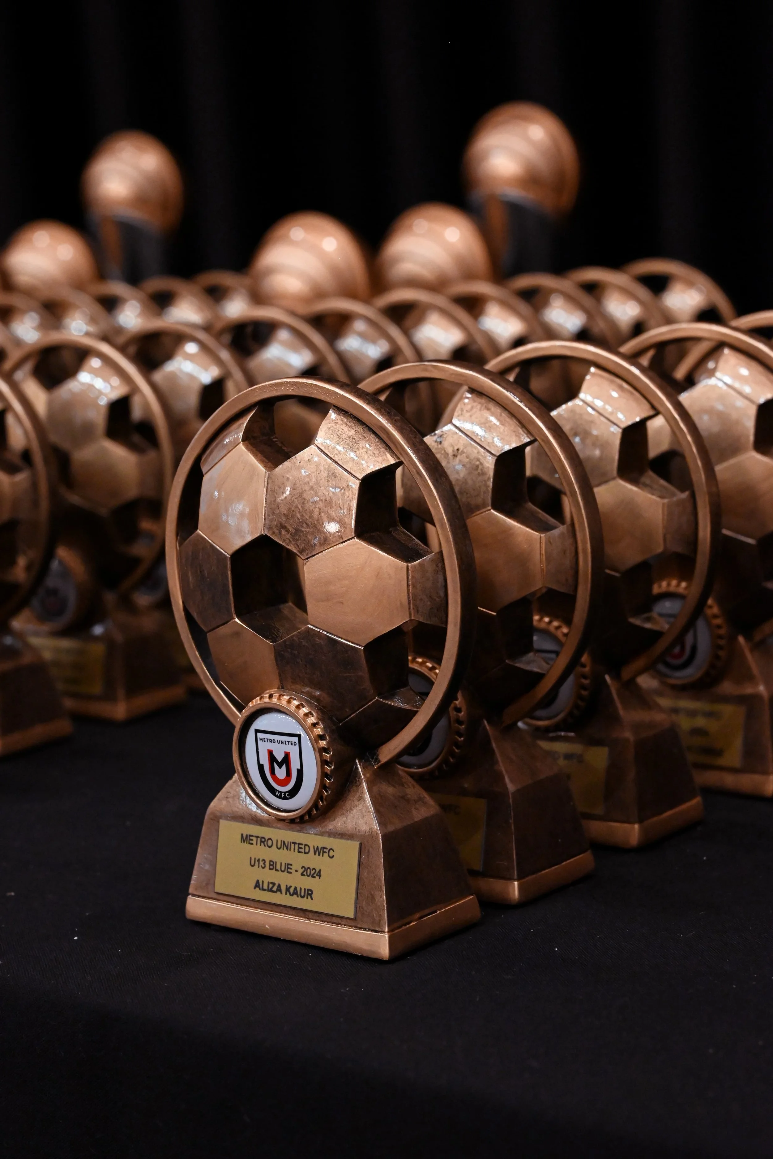 Multiple bronze soccer trophies with a black background and a gold plaque that reads 'METRO UNITED WFC U13 BLUE - 2024 ALIZA KAUR' celebrated at Donato the venue in Adelaide