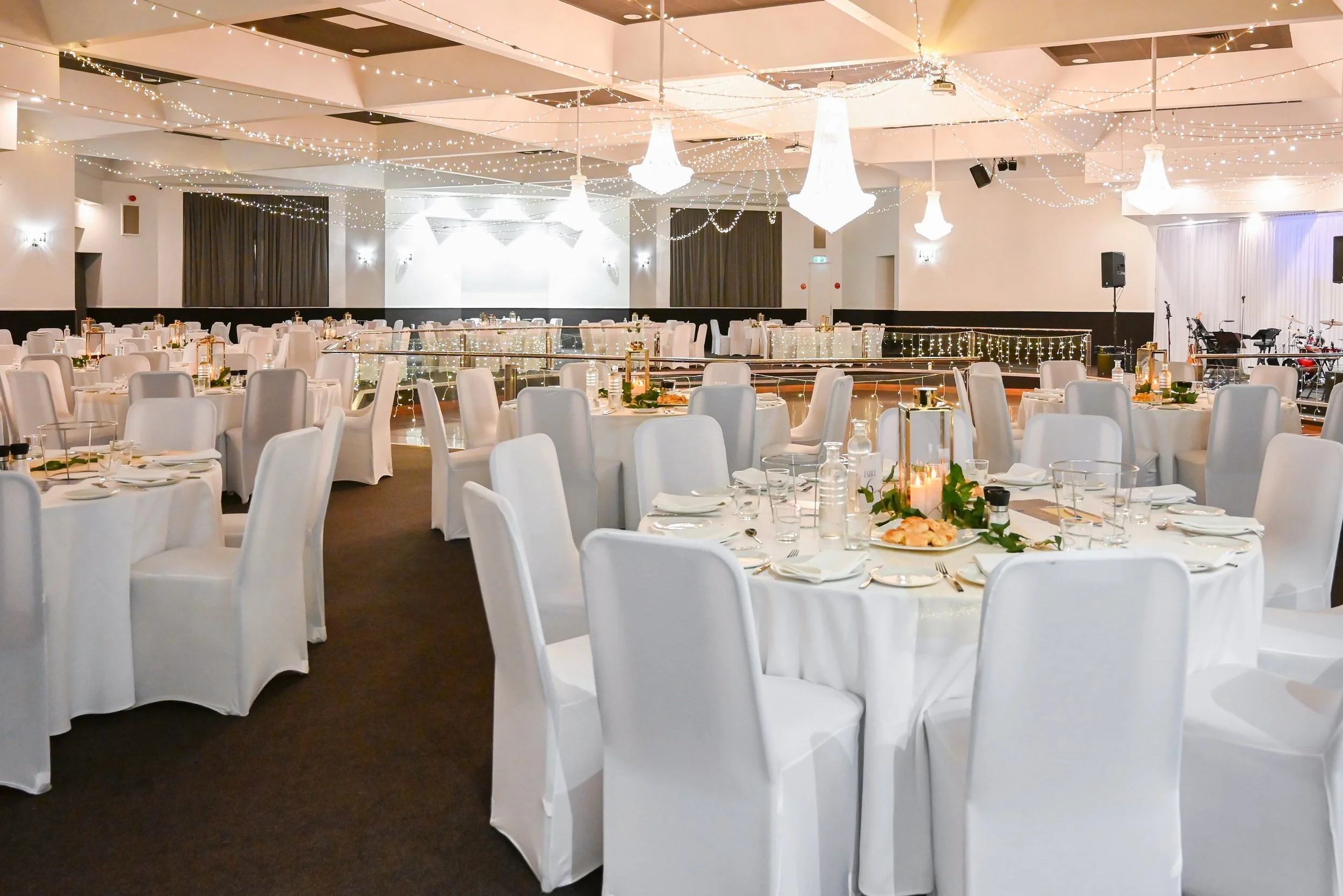 Ballroom decorated for an event with white tablecloths and chairs, string lights, chandeliers, and a small stage with musical instruments.