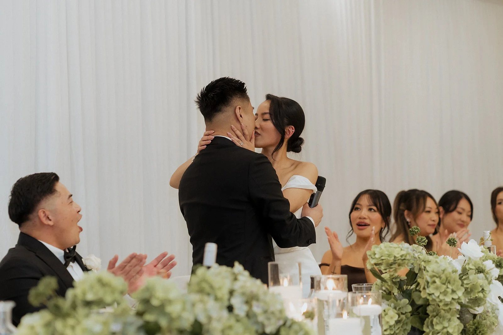 A bride and groom share a kiss during their wedding reception while guests claps and cheer around them, with decorative flowers on the table at Donato reception centre in Adelaide