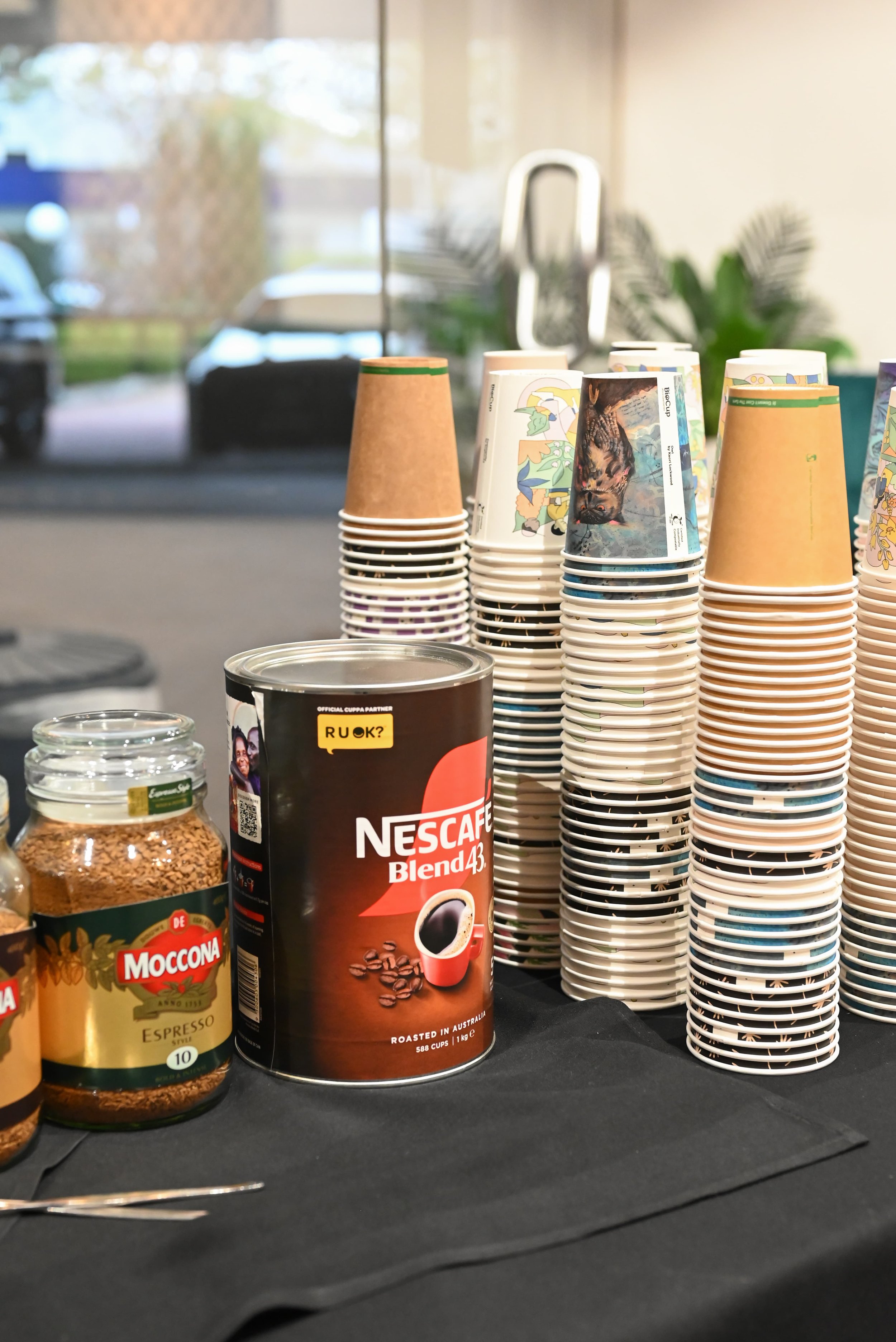 Stacked paper coffee cups with various designs, a can of Nescafe Blend 43 coffee, and a jar of Moccona coffee on a black surface near a window with parked cars outside.