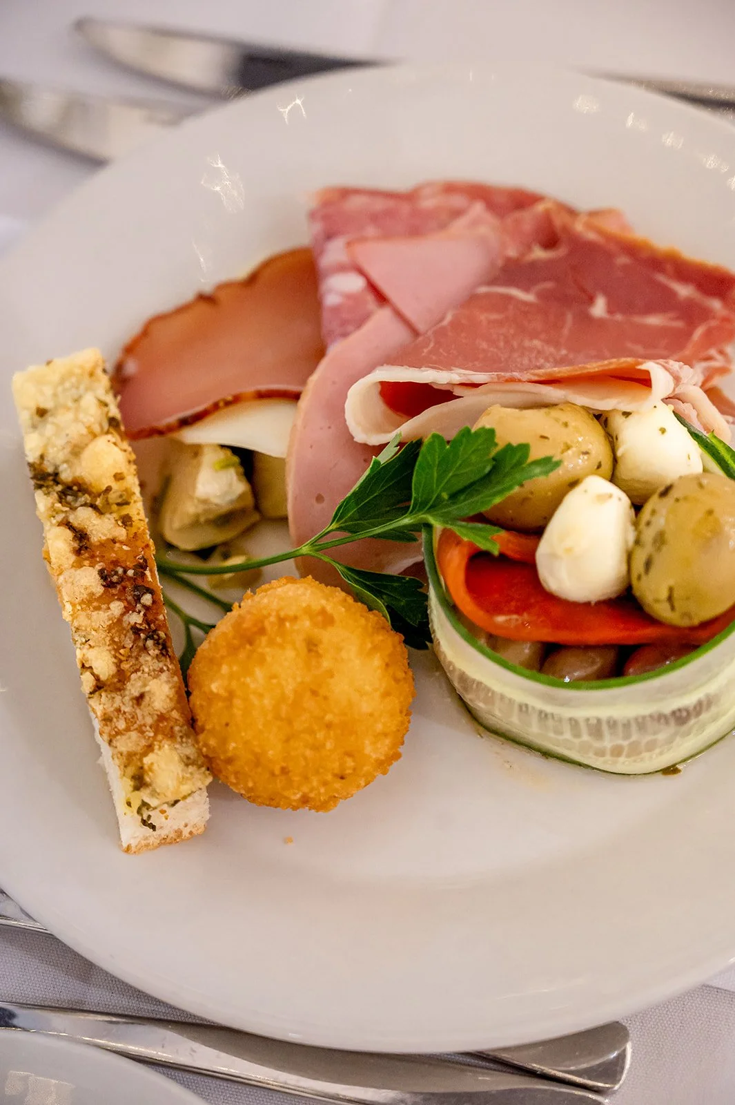 Assorted appetizers on a white plate, including slices of cured meats, cheese with herbs, marinated olives, a breadstick, and a croquette garnished with parsley.