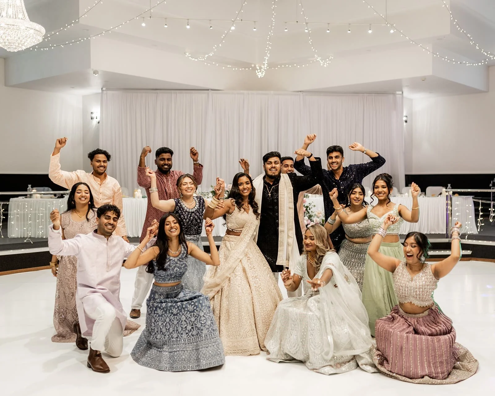 A group of people celebrating at a wedding reception at Donato the venue, some wearing traditional Indian clothing, dancing, and smiling inside a decorated hall with string lights and a white backdrop.