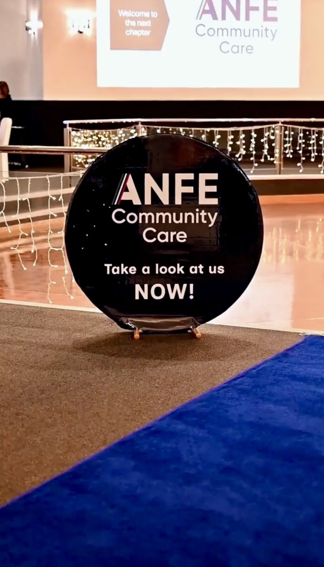 Black circular sign with white and pink text reading "ANFE Community Care Take a look at us NOW!" placed on a hallway floor. In the background, there is a decorated area with string lights and a large screen displaying a welcome message for the next 