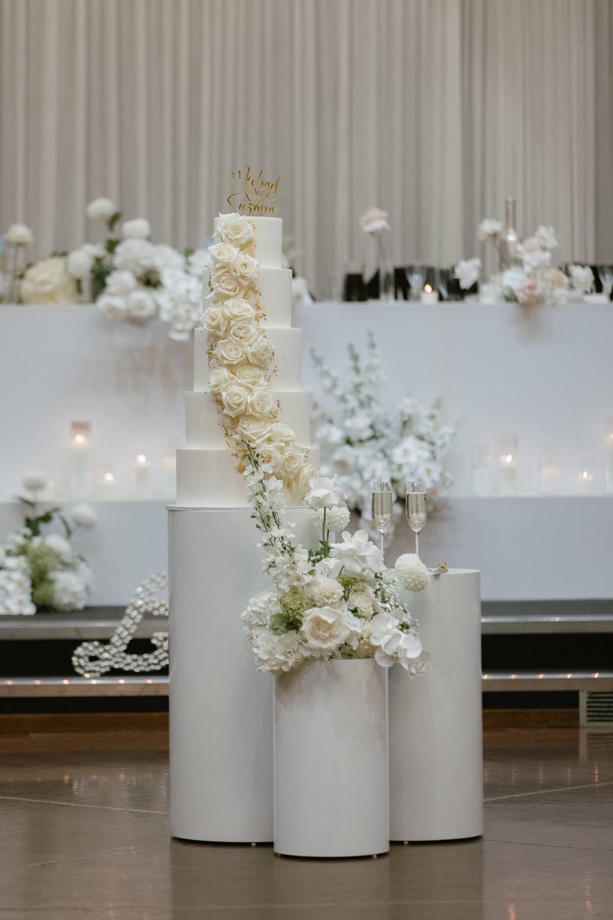 Elegant wedding cake with cascading white roses and flowers, situated on white cylindrical pedestals, with a backdrop of white floral arrangements and drapes at Donato the venue in Adelaide