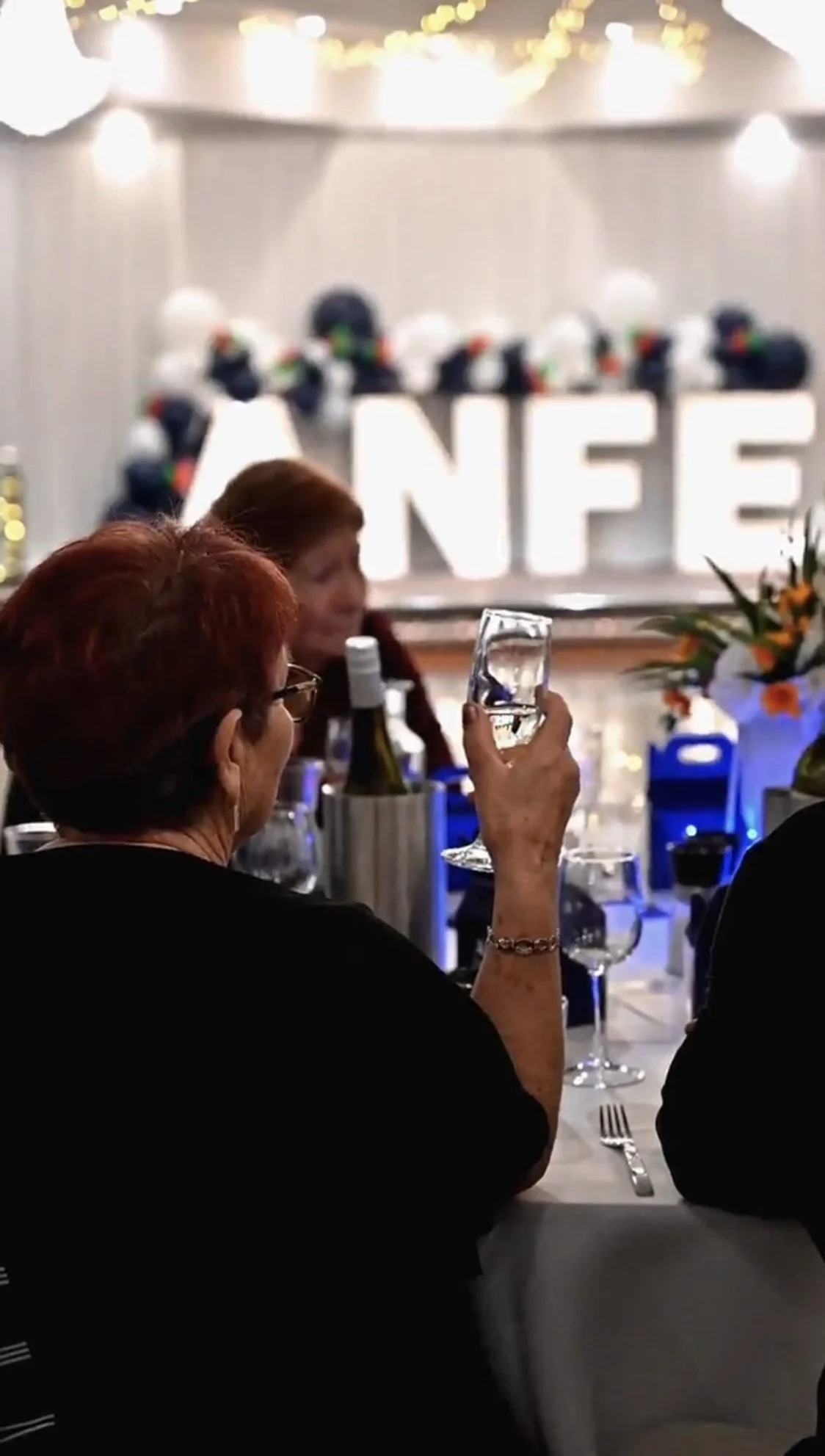 Guests seated at a table taking a photo of a large decorated cake with the letters 'NFE' in the background, surrounded by string lights and floral arrangements.