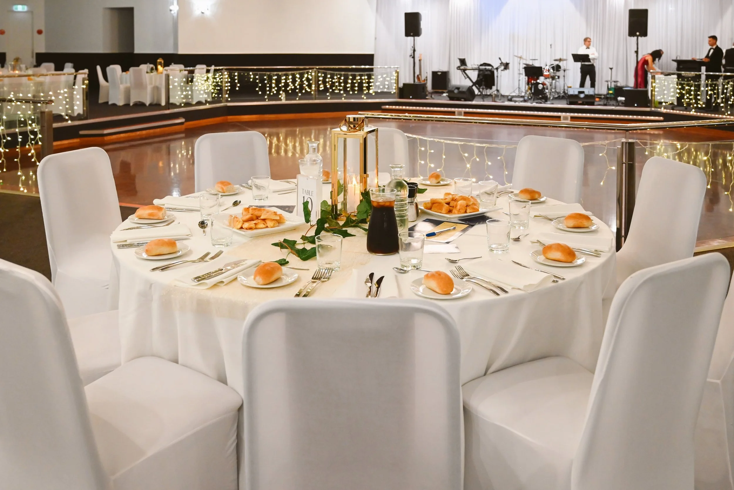 Round banquet table set with white chairs, plates, bread rolls, glasses, and silverware, with a small green plant and candle centerpiece, in a decorated event hall with a stage and musical instruments in the background.
