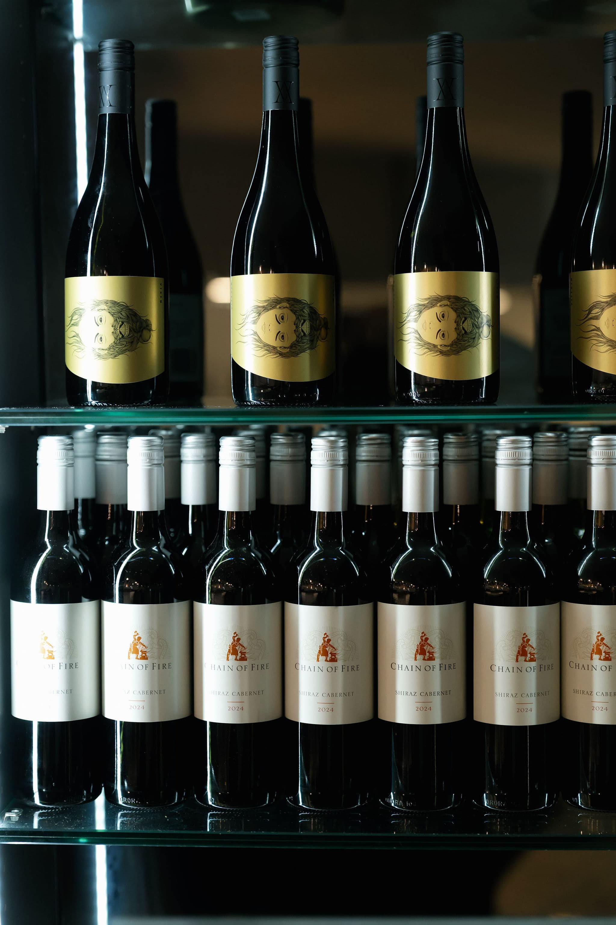 Bottles of wine on a glass shelf behind a glass door. The top row has bottles with a yellow label featuring a face illustration, and the bottom row has bottles with a white label and a logo of a horse and rider.