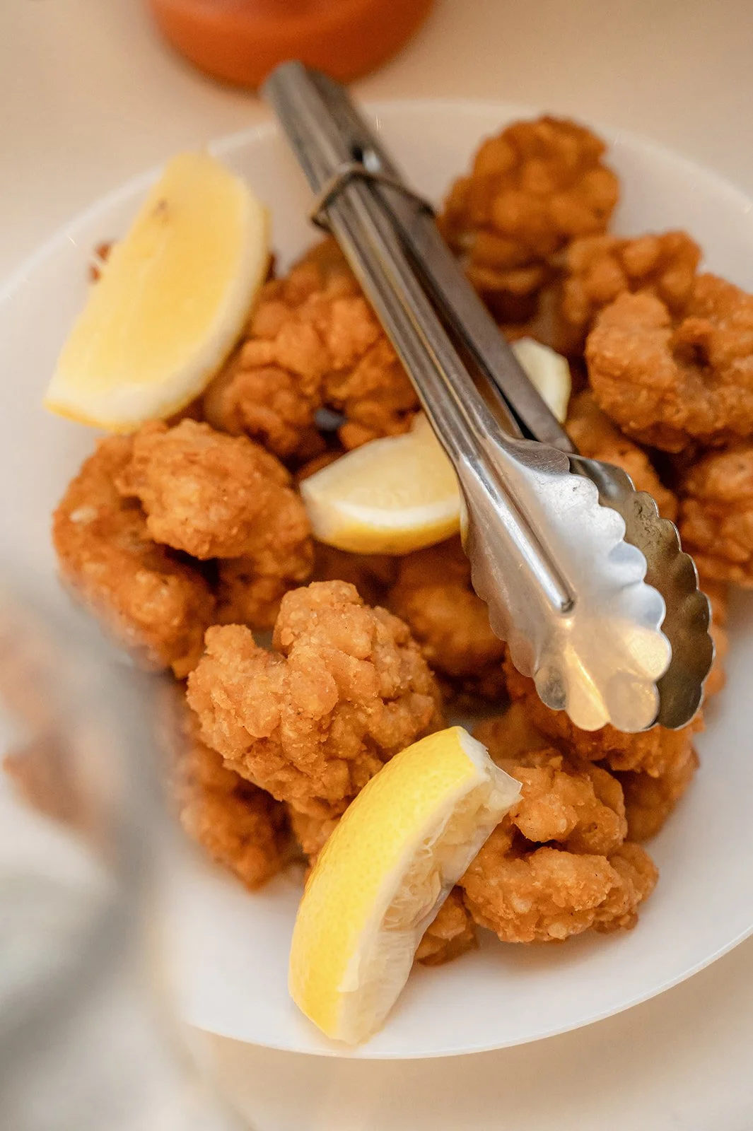 Fried chicken bites with lemon wedges and metal tongs in a white bowl.