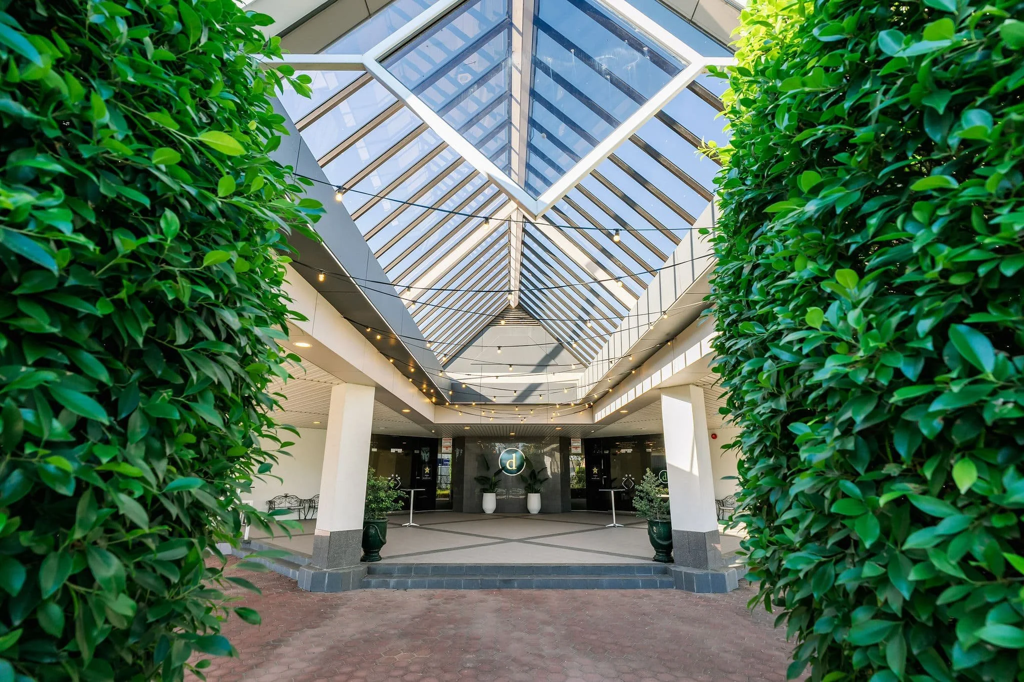 Entrance of a shopping mall or commercial building with greenery on both sides and a glass skylight ceiling overhead.