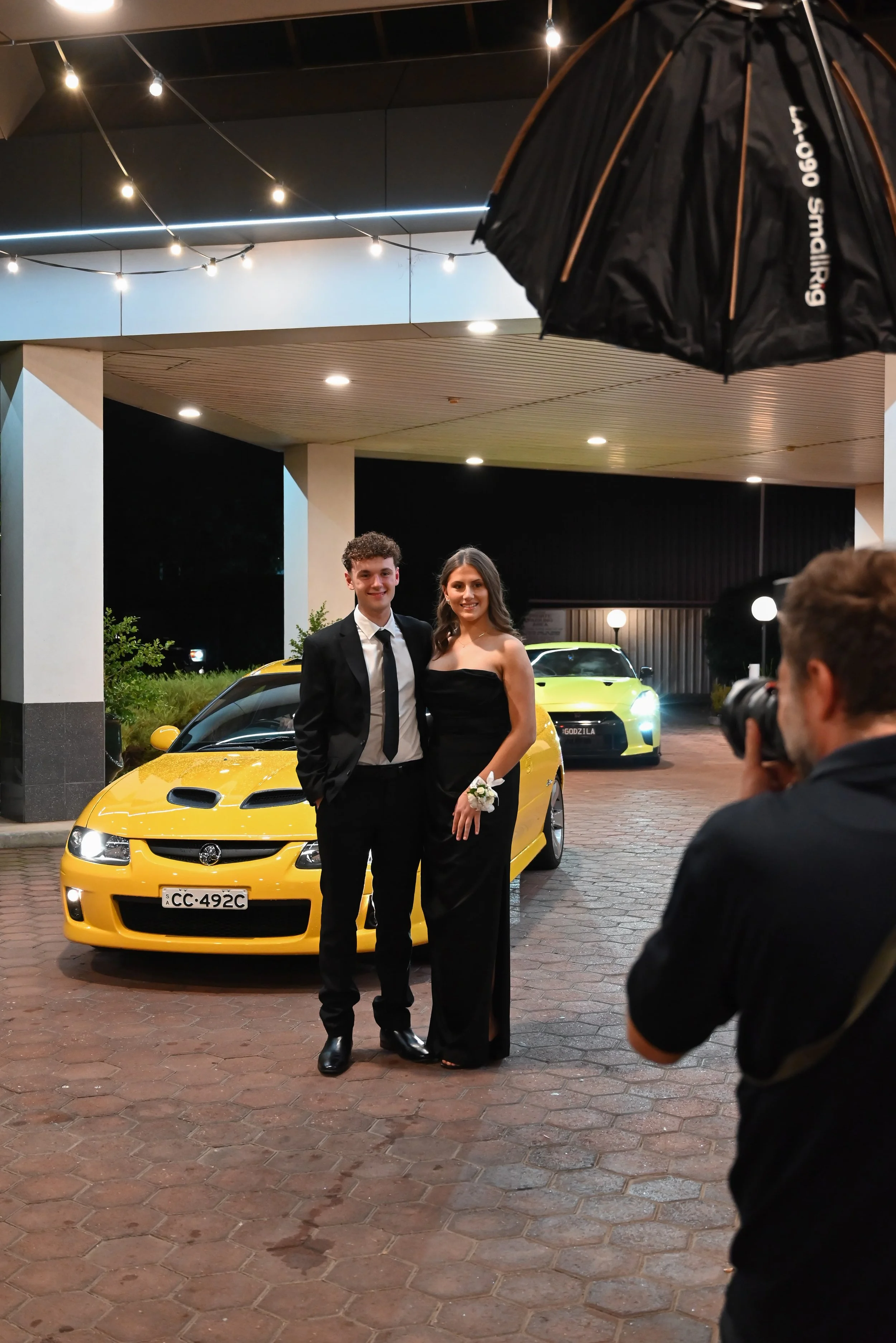 A young couple dressed in formal attire, standing in front of a yellow sports car, during a nighttime event. The man is wearing a black suit with a white shirt and black tie, and the woman is in a black strapless gown, holding a small bouquet. A phot