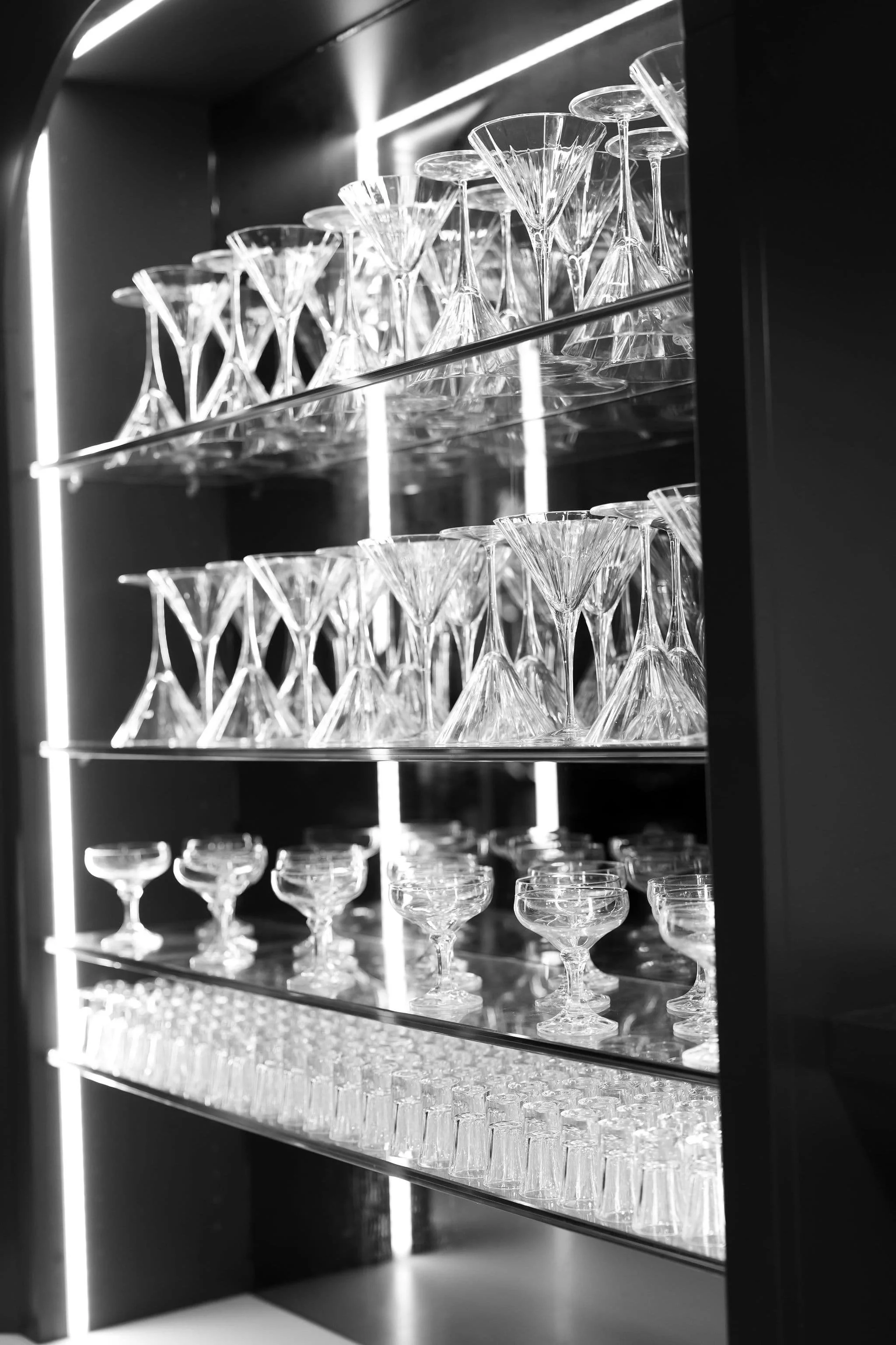 Multiple empty elegant glassware including cocktail glasses and martini glasses arranged on glass shelves in a display case, with a modern black and white aesthetic.