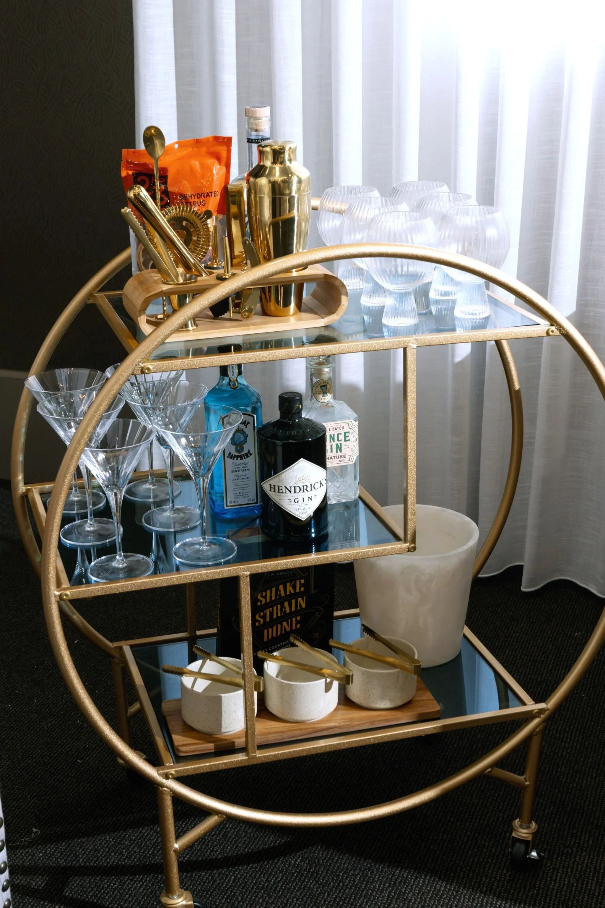 A gold bar cart holds glassware, spirits, and bar accessories, with a white curtain in the background.