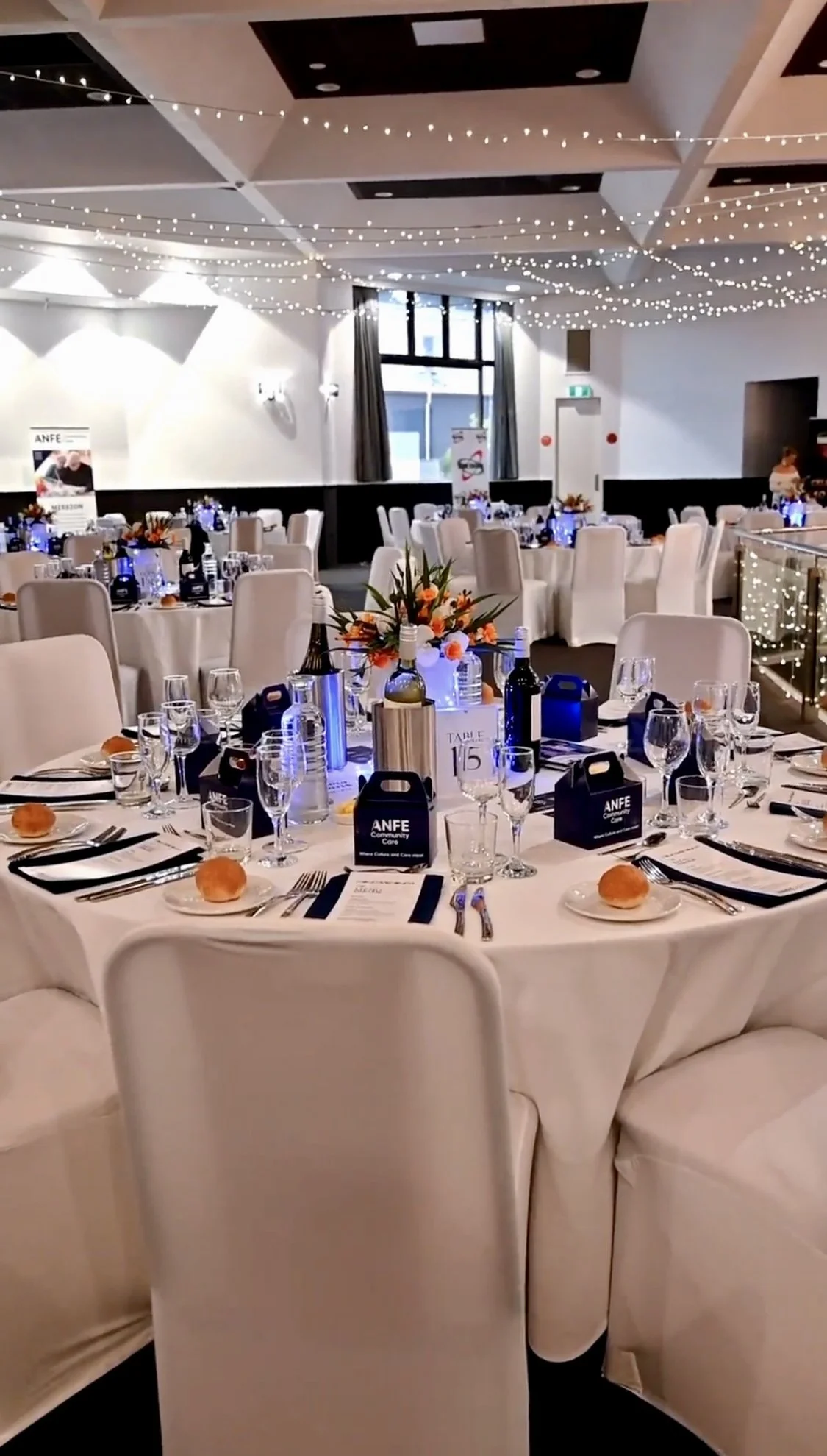 An elegantly decorated banquet hall set for a formal event, with round tables covered in white tablecloths, floral centerpieces, wine bottles, glassware, and black takeout boxes labeled 'ANFE Community Care.' The hall features string lights hanging f