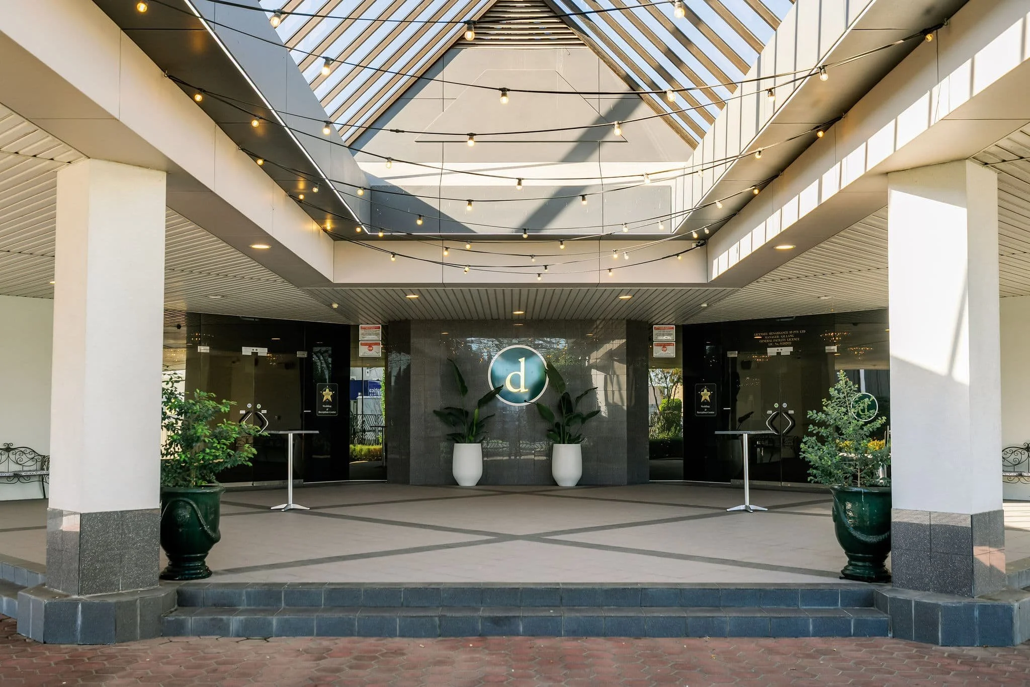 The entrance of a modern building with a glass roof and outdoor string lights. There are two large white planters with greenery, a black wall with signages, and glass doors, with potted plants on either side.