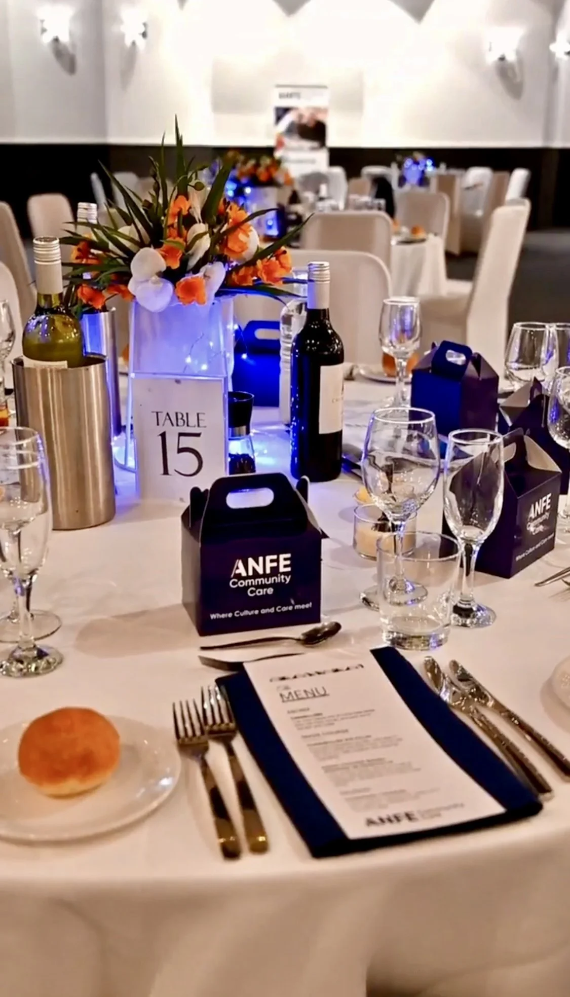 A banquet table with a centerpiece of orange and white flowers, surrounded by wine bottles, glasses, silverware, and black gift bags labeled 'ANFE Community Care.' The table has a card with ‘Table 15’ written on it, and a menu is placed at each seat.