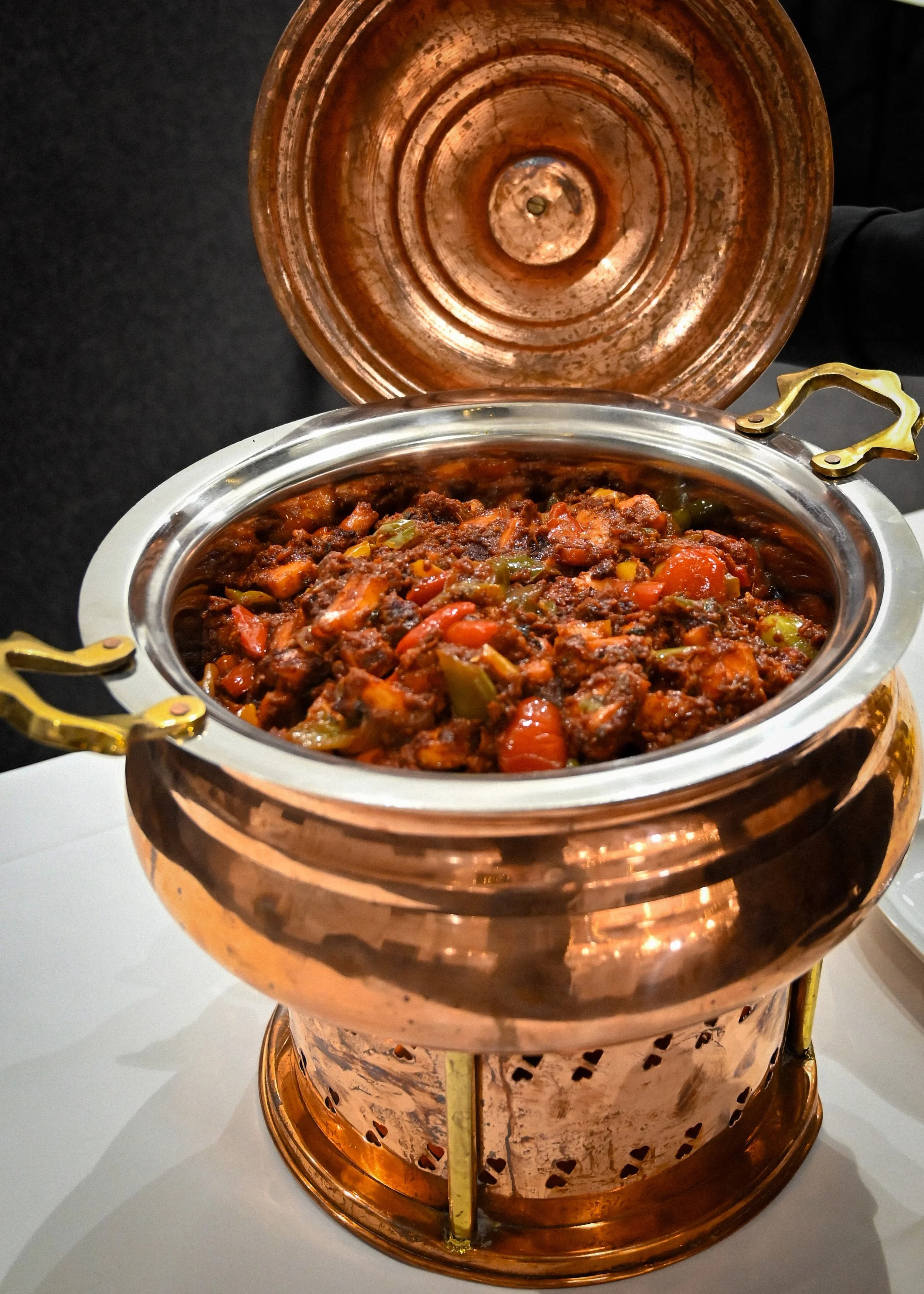 A copper food warmer with a lid open, revealing cooked stew with vegetables inside, placed on a white surface.