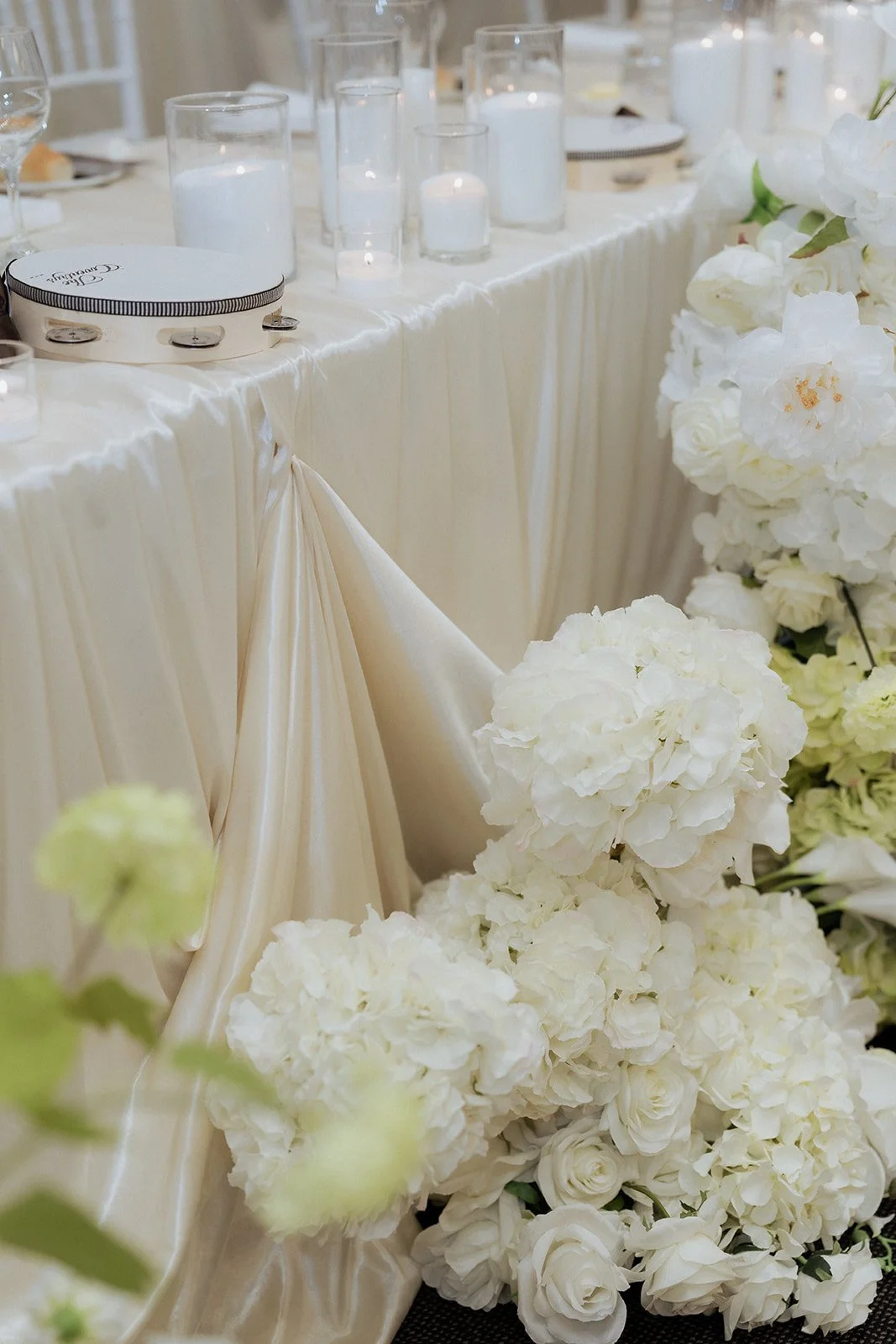 A decorated table with white flowers, candles in glass holders, and a white satin draped tablecloth, likely at a wedding or formal event.