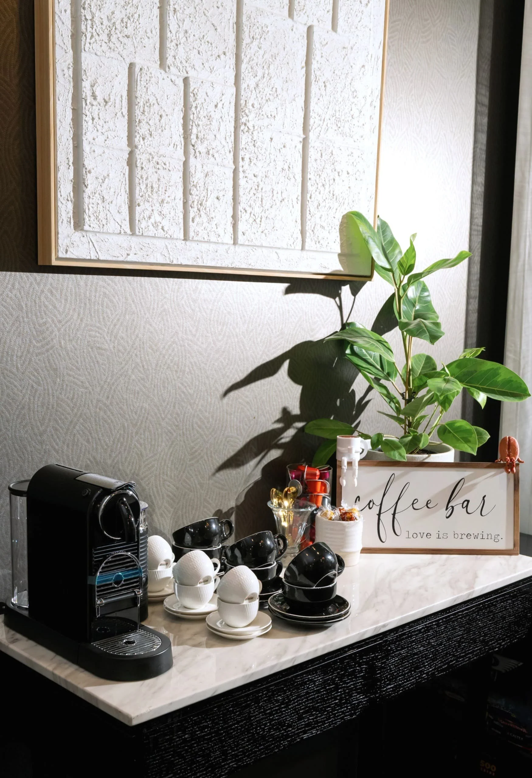 Coffee bar setup with white and black cups, a coffee machine, and a decorative sign that reads 'coffee bar love is brewing.' A green plant is also visible on the counter.