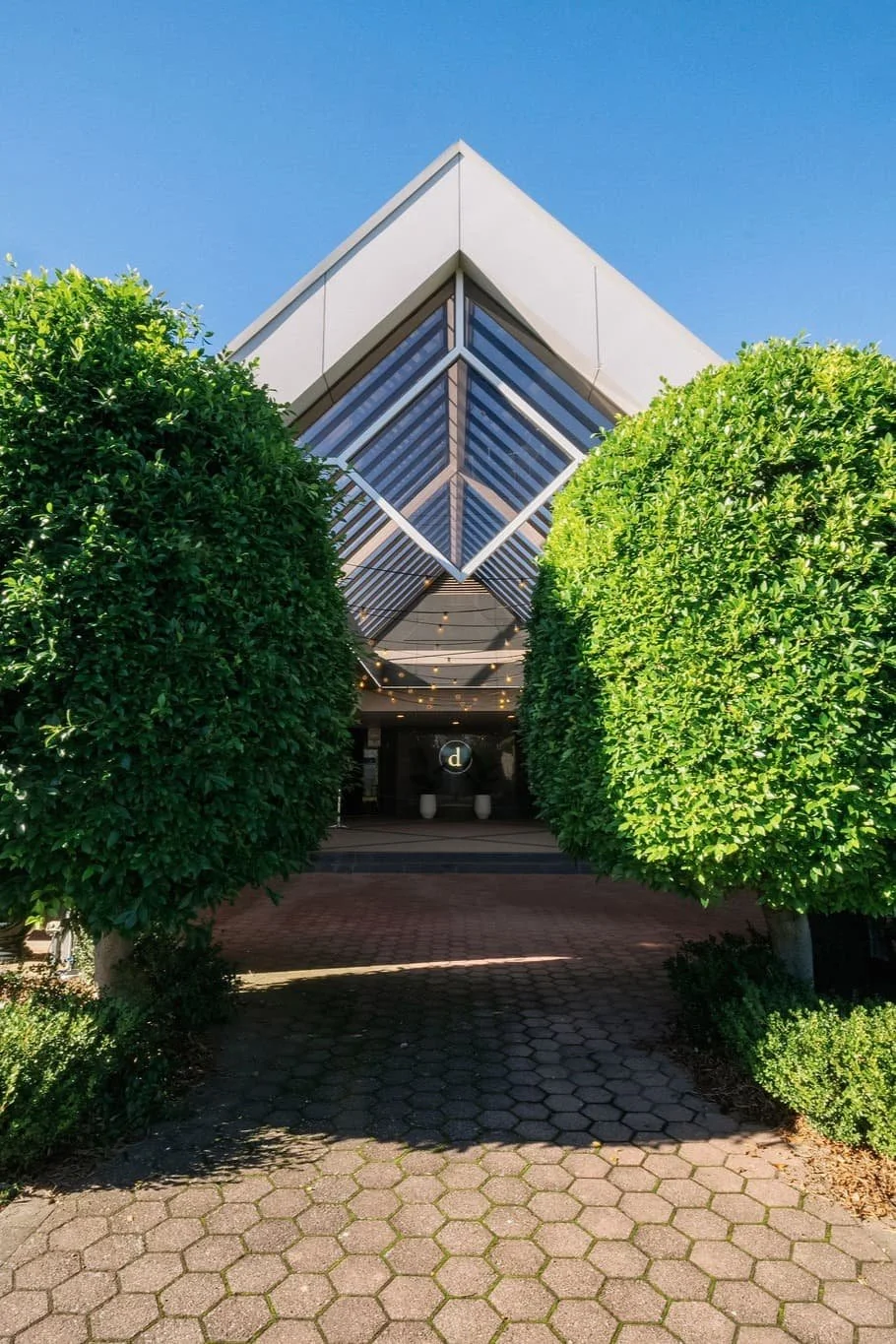 Modern building with an angular white roof and glass windows, framed by two large, rounded green bushes in the foreground.