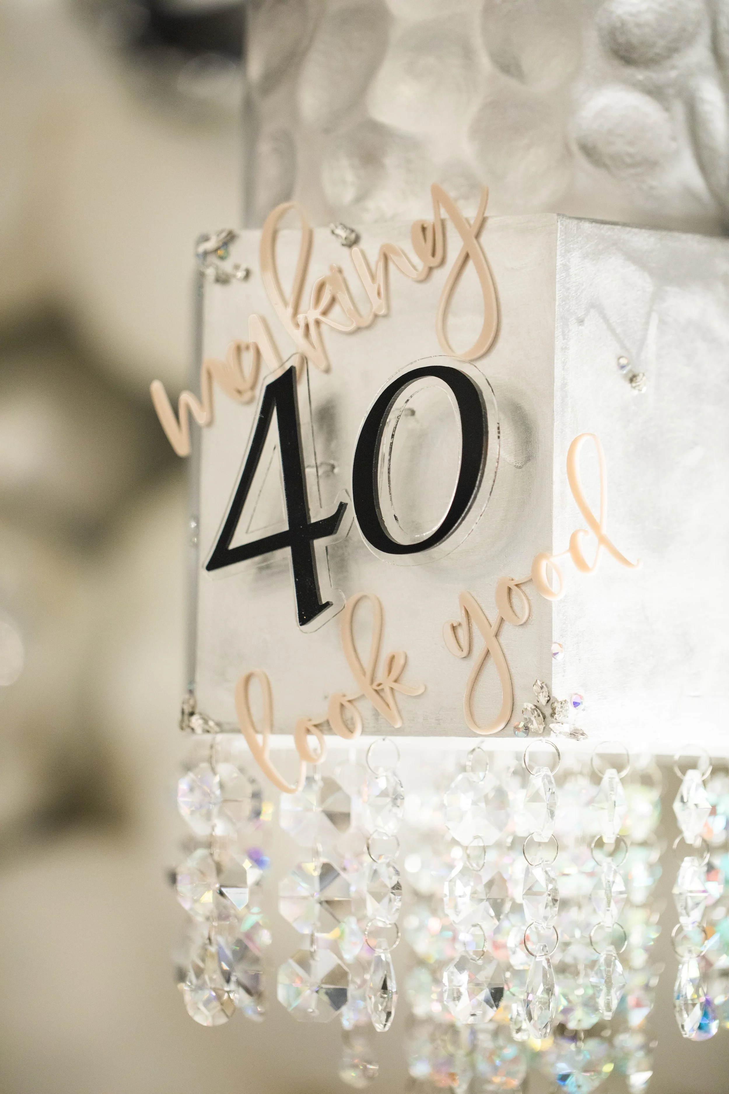 Close-up of a decorative hanging sign that reads 'happy 40th' with a crystal chandelier in the background.