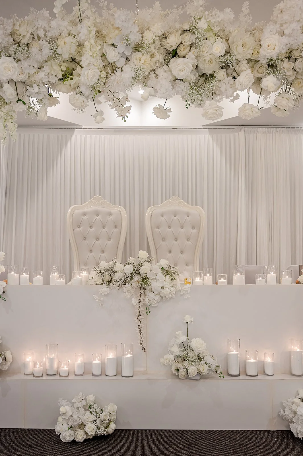 Elegant white wedding reception table with two ornate white high-back chairs, surrounded by white floral arrangements and many white candles in glass holders, with a white draped backdrop and a floral ceiling installation at Donato reception centre 