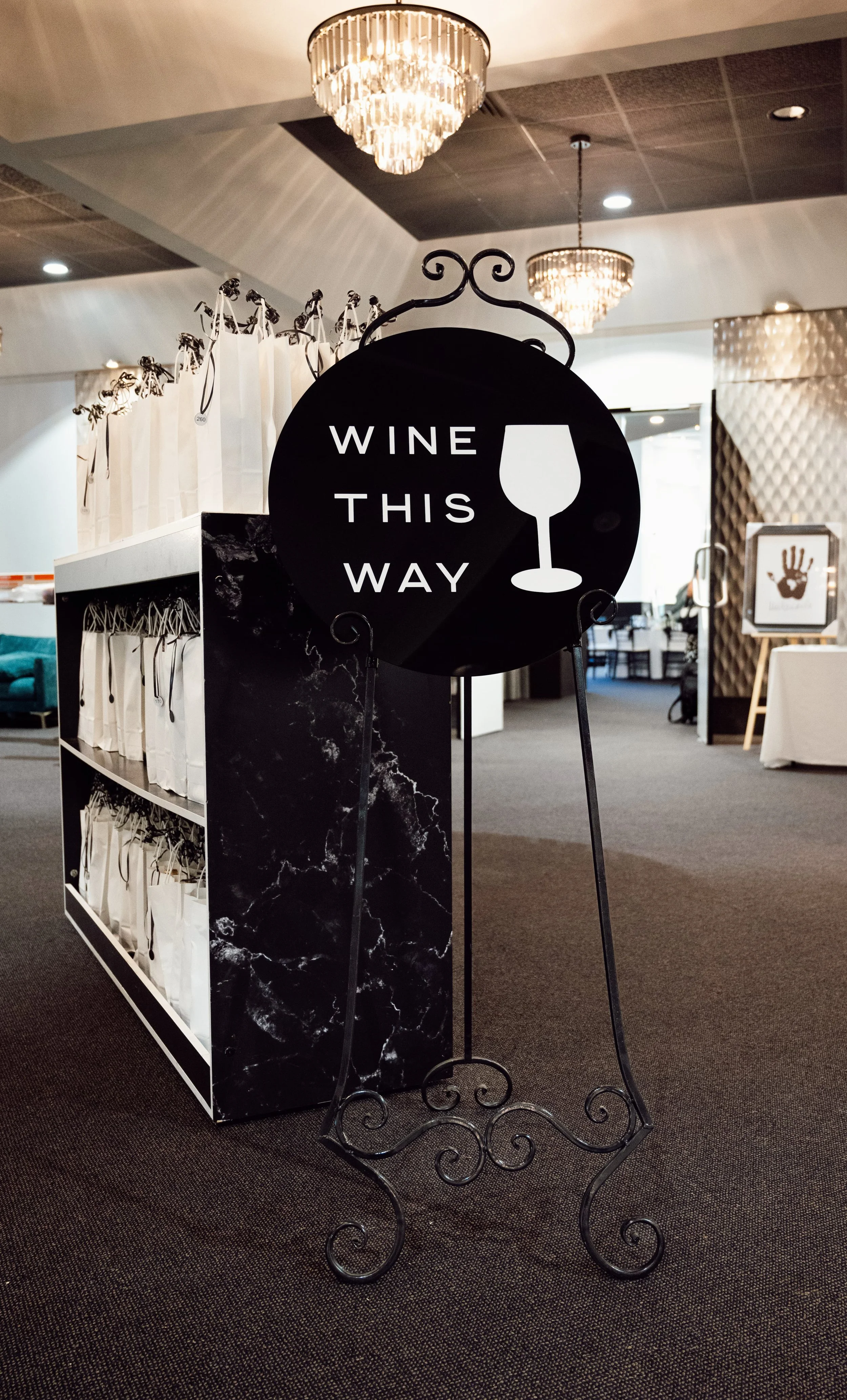 A black circular sign with the words "WINE THIS WAY" and a wine glass icon, situated on a decorative black wrought-iron stand in a modern indoor space, possibly a wine shop or event area.