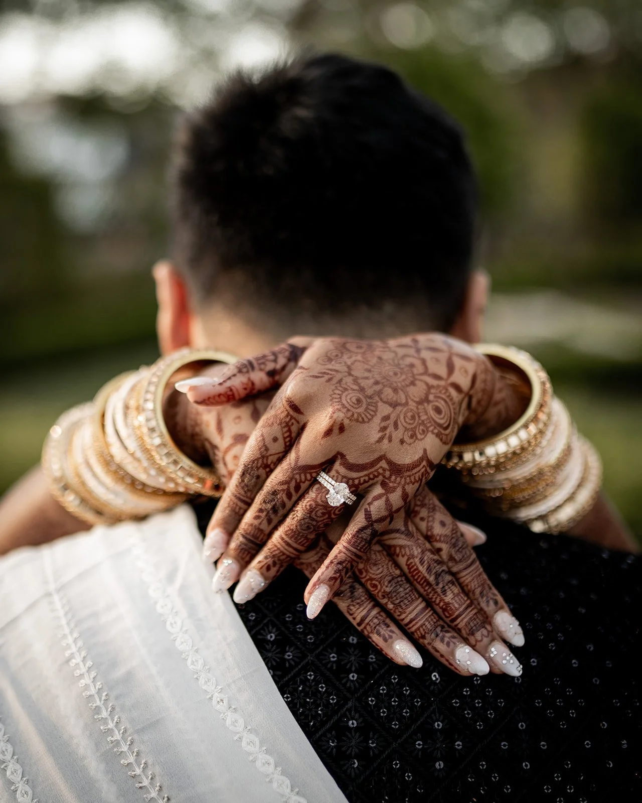 A close-up of a person with mehndi (henna) decorated hands and wearing multiple gold bangles, rings, and jewelry. The person is resting their hands on another person’s shoulder, who is wearing a white embroidered garment. The background is blurred wi