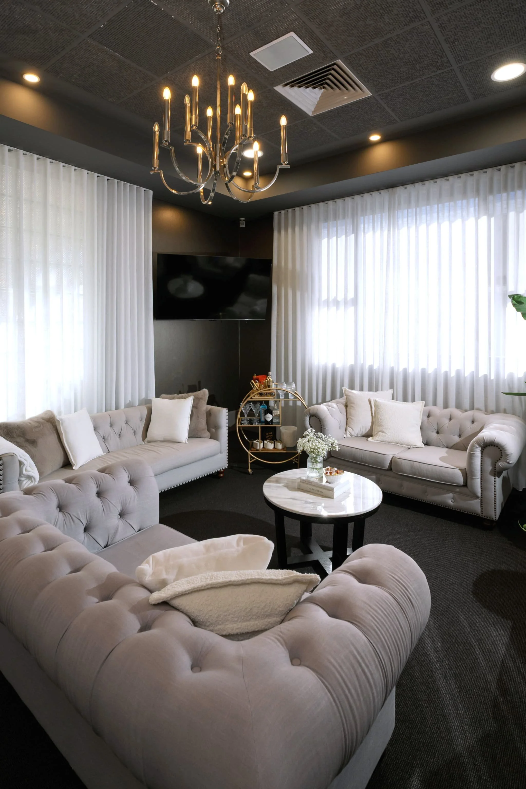 A luxurious living room with tufted beige sofas arranged in a semicircle, adorned with plush pillows. A small round marble coffee table with a floral arrangement and books is in the middle. White curtains cover large windows, a black TV is mounted on