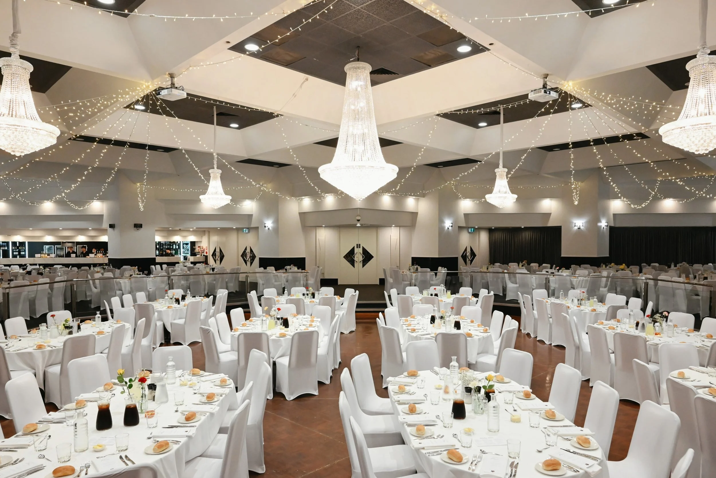A banquet hall decorated with white tablecloths and chairs, adorned with floral centerpieces, glass bottles, and place settings. The ceiling features elegant chandeliers and string lights, creating a festive ambiance for an event or celebration.