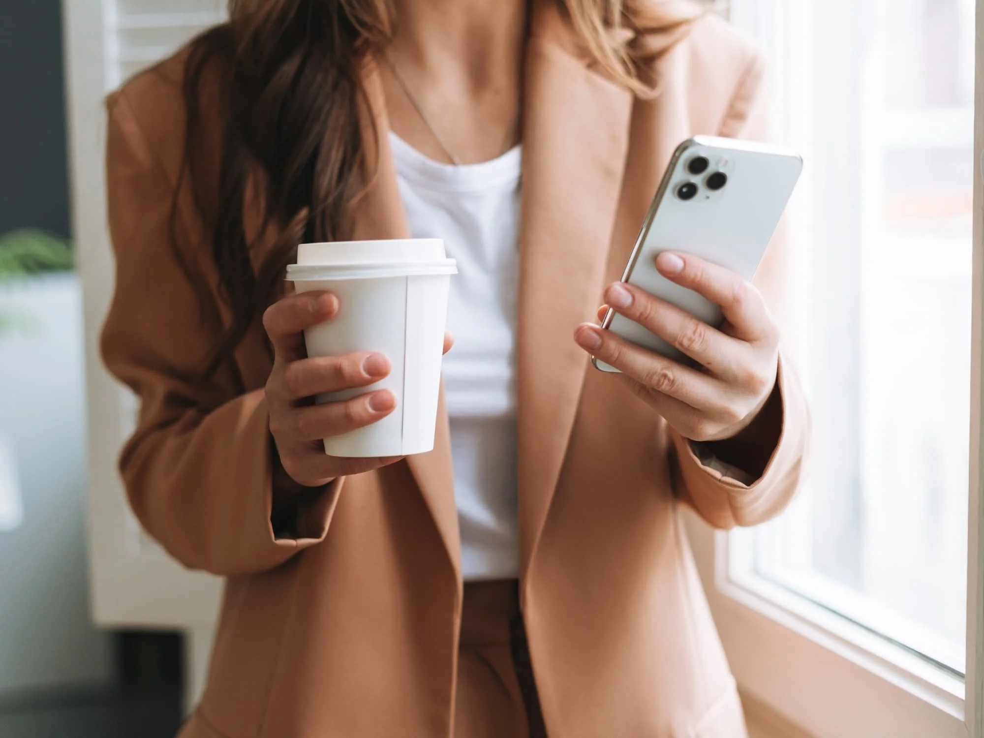Woman holding a white disposable coffee cup and looking at her smartphone near a window in a sunlit room.