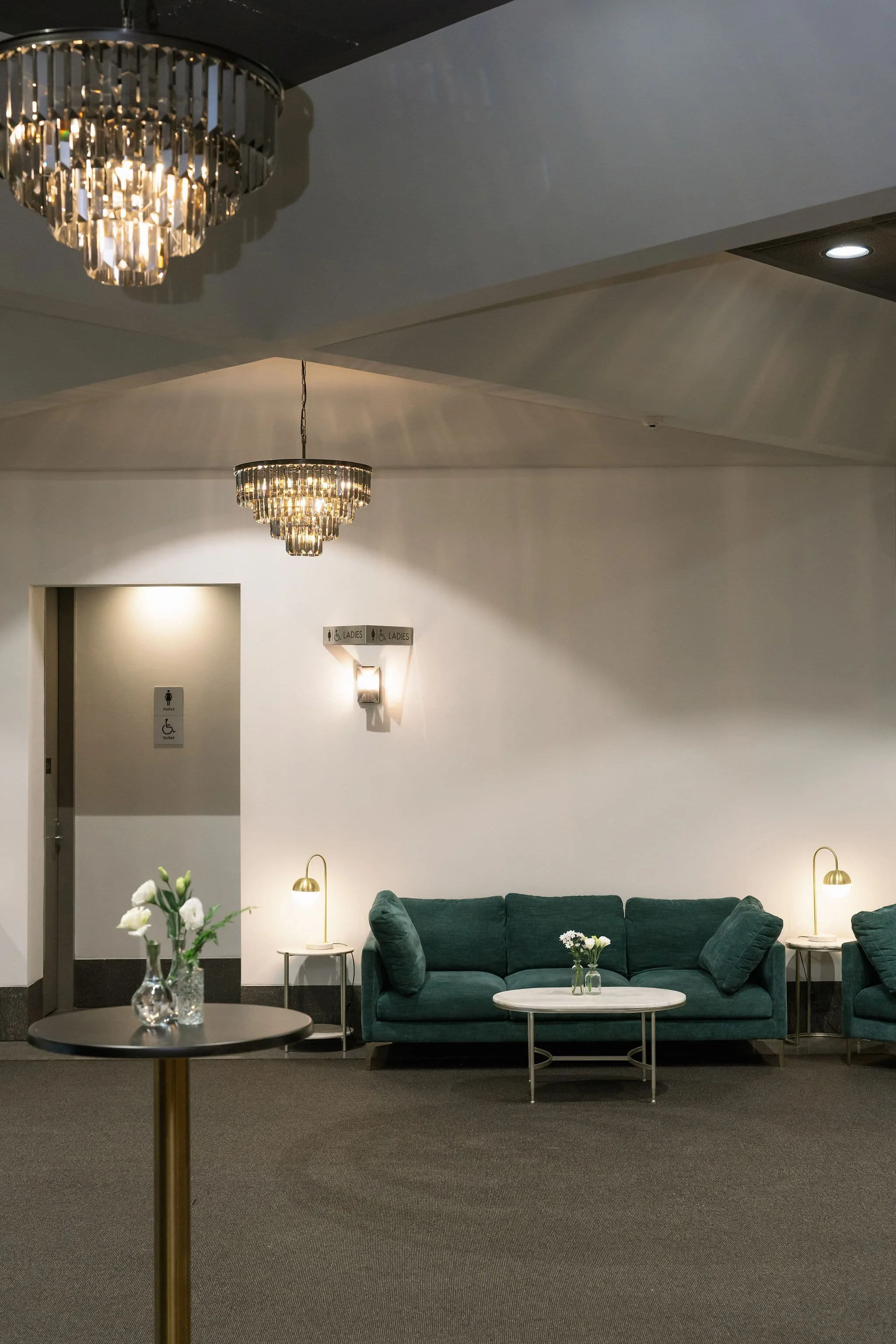 A hallway with a dark carpet, a green sofa, and white walls. Two gold and white lamps on side tables, a coffee table with a flower arrangement, a chandelier, and a sign indicating women and accessible restrooms.
