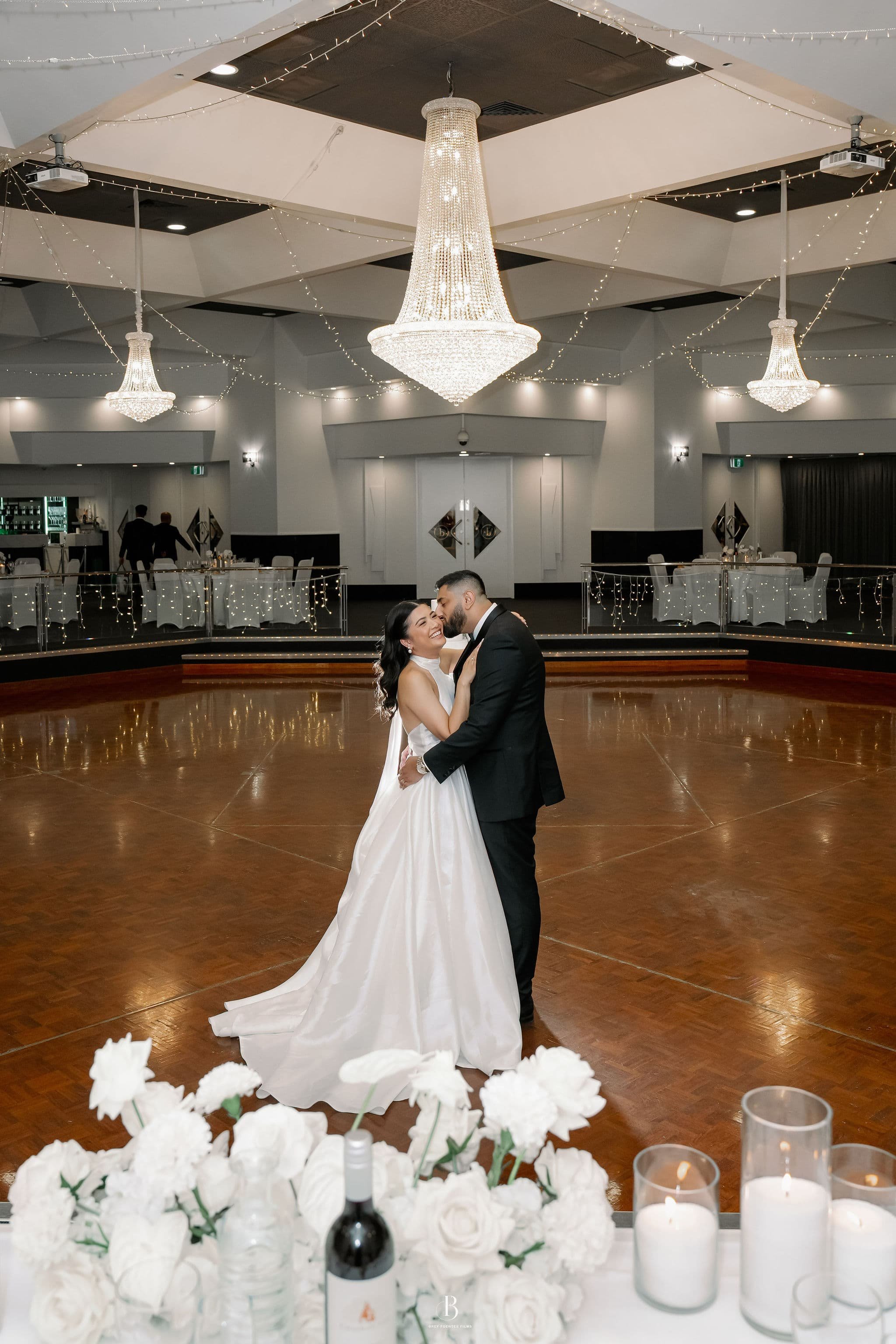 A bride and groom are dancing in a decorated reception hall with elegant chandeliers and string lights. The bride is wearing a white wedding gown, and the groom is in a black suit. The foreground has a table with white roses, candles, and a wine bott