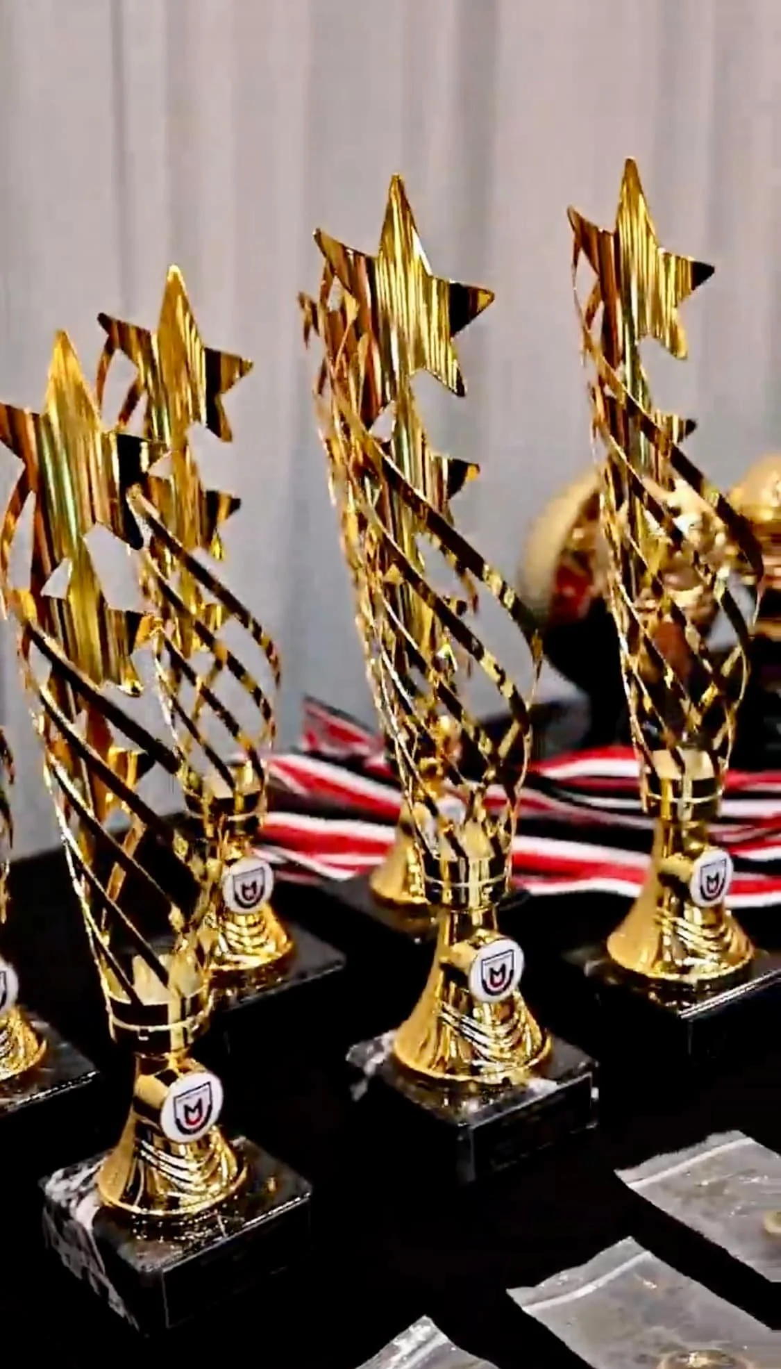 Multiple gold-colored trophies with a black base arranged on a table, with some medals and striped cloth visible in the background.