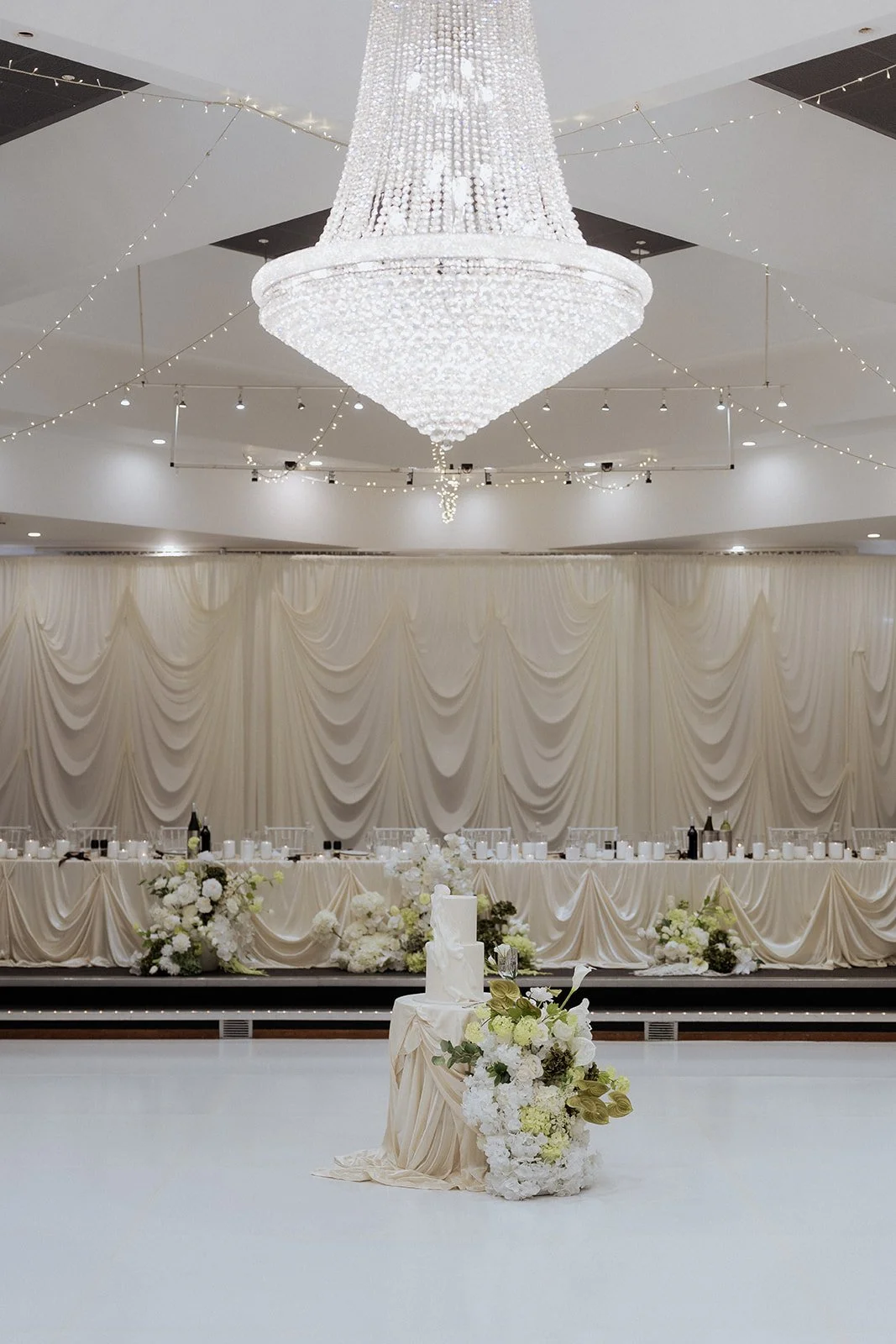 Elegant wedding reception hall with a grand chandelier, draped cream curtains, floral arrangements, and a long table for guests.