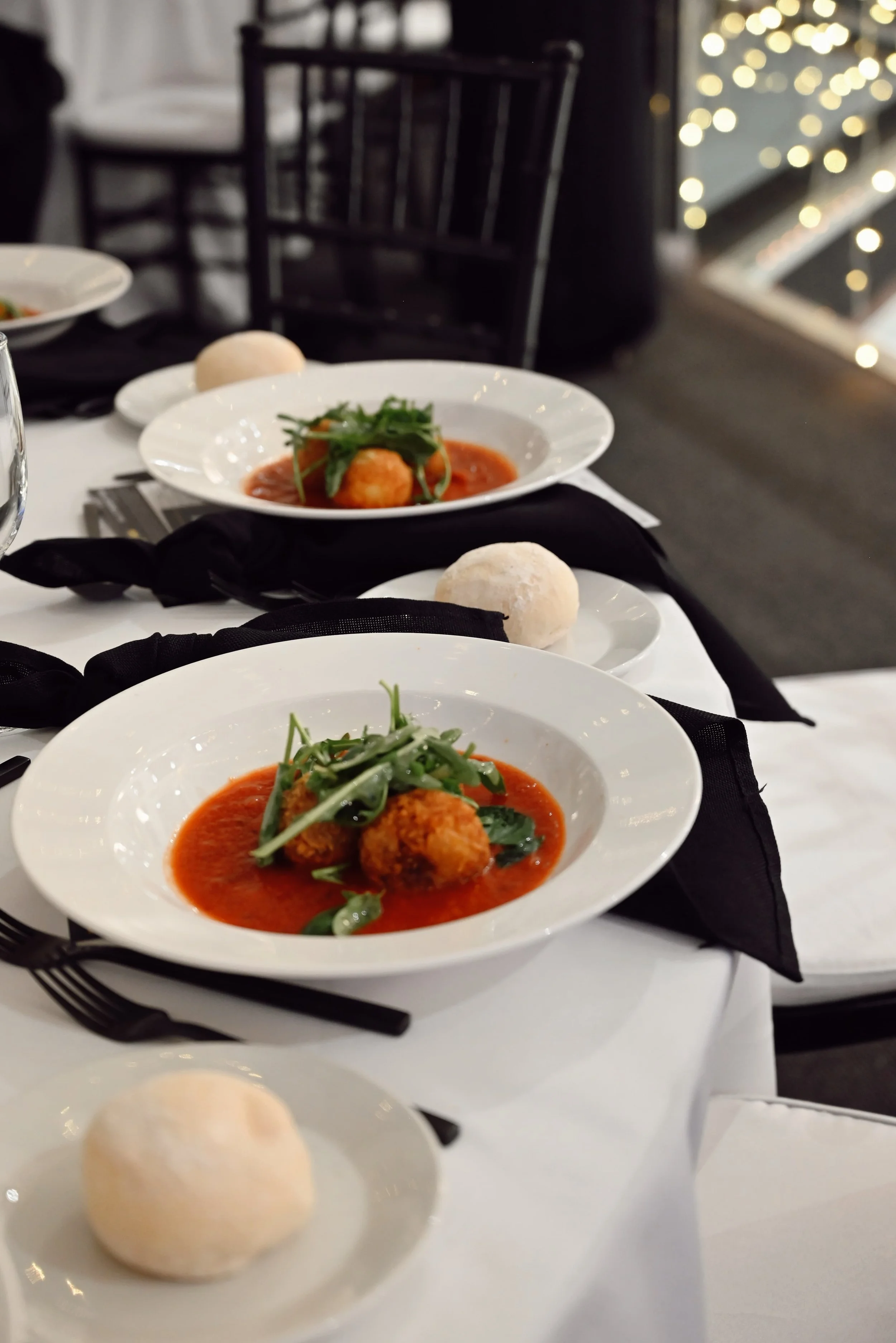 White plate with a breaded appetizer topped with greens in red sauce, on a white table with black utensils and a black napkin, blurred background of restaurant setting with lights.
