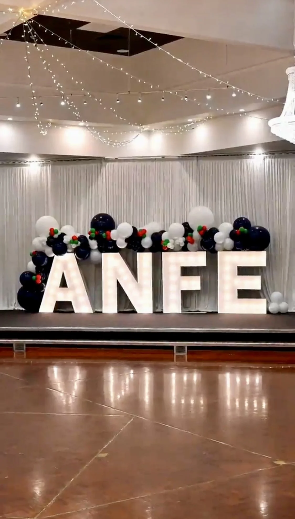 Decorative birthday display with large illuminated letters spelling 'ANE' and a balloon arrangement in black, white, and red, with string lights and tinsel in the background.