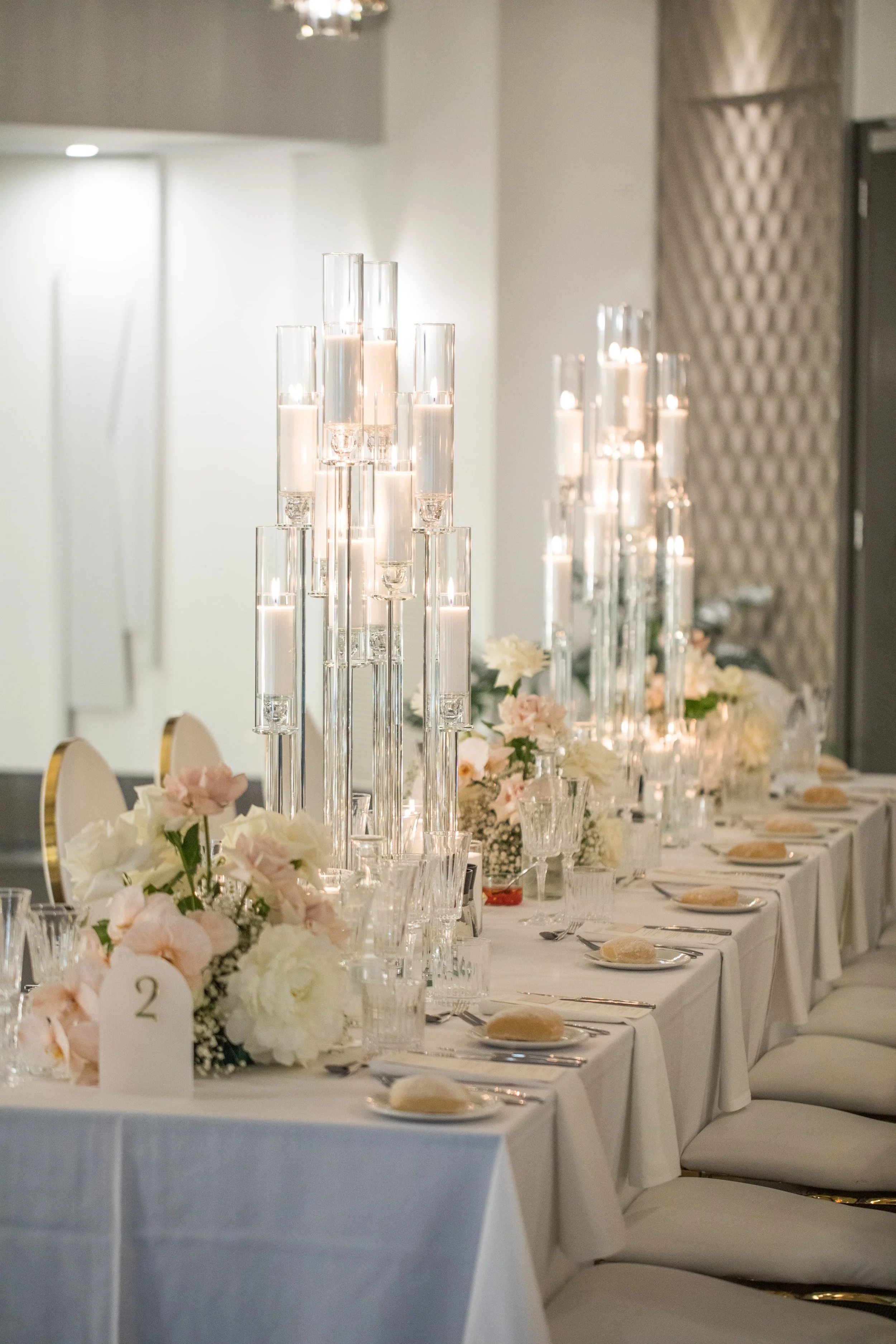 Elegant banquet table setup with tall glass candle holders, floral arrangements, and place settings with bread rolls and utensils, in a well-lit reception hall.