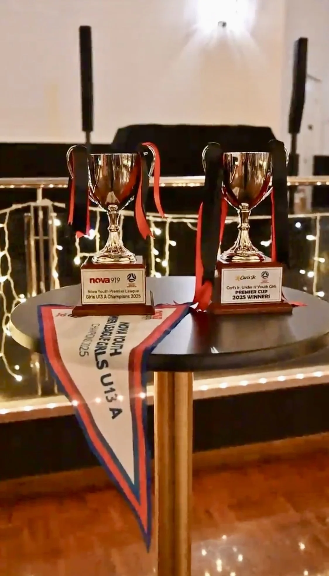 Two trophies on a table, labeled for youth rugby league championships, with a ribbon and a banner in the background, in a festive indoor setting.