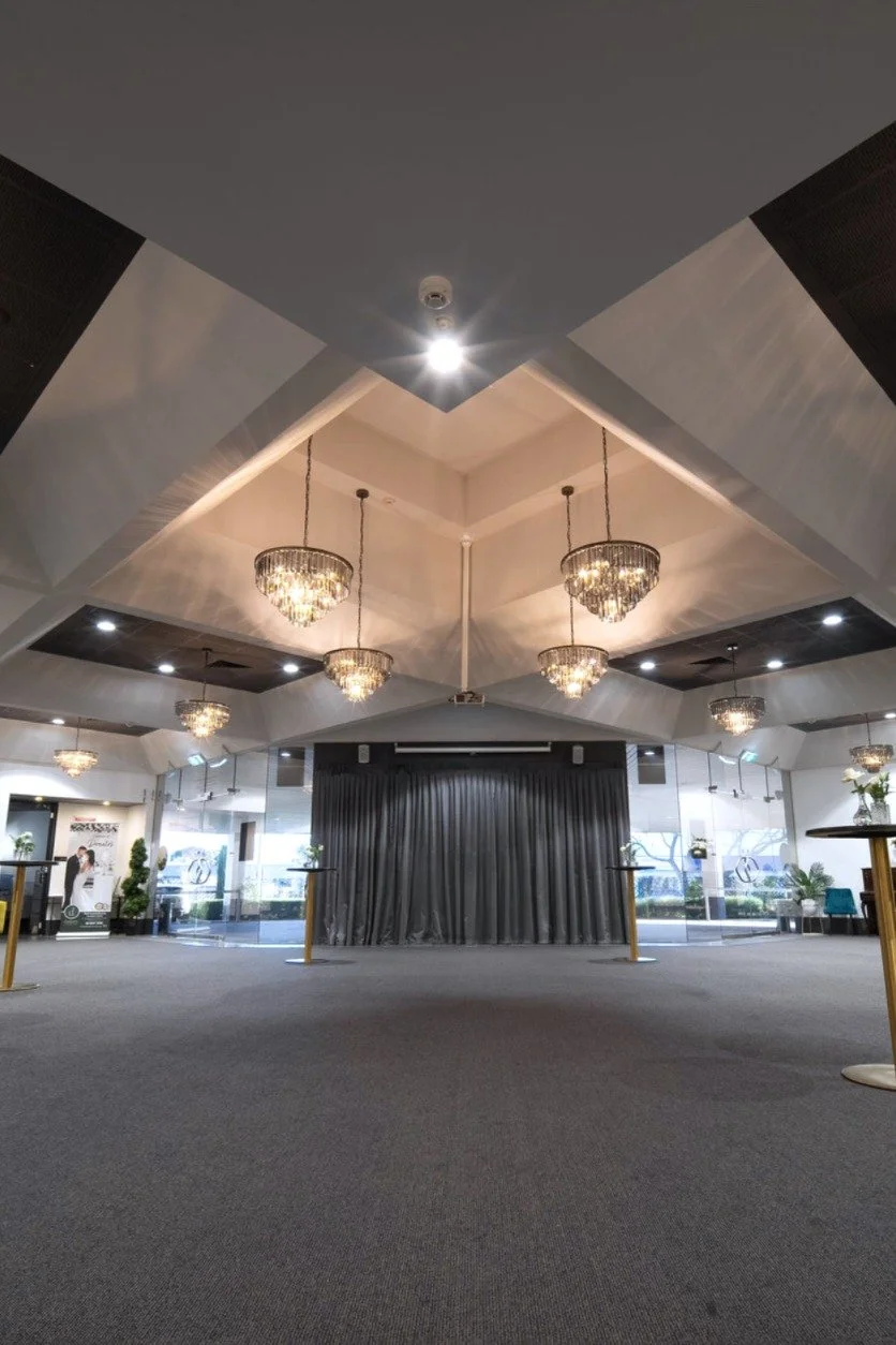 Empty event or conference room with chandeliers and a stage with a black curtain, large windows, and seating areas.