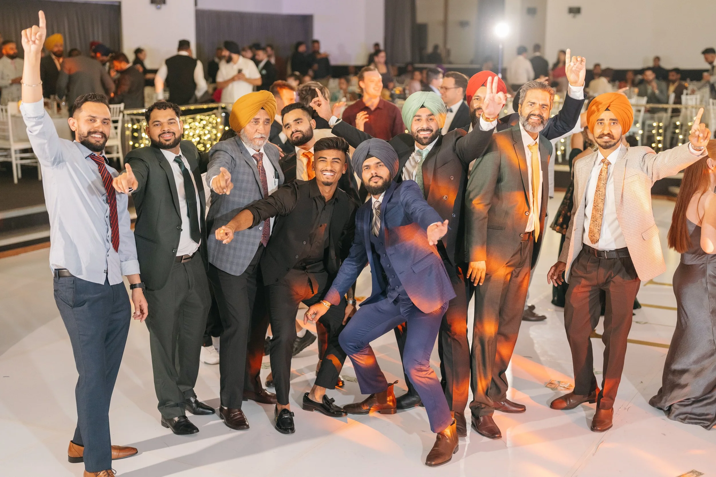 Group of men in suits and traditional Indian turbans dancing and celebrating on a dance floor at a wedding reception.