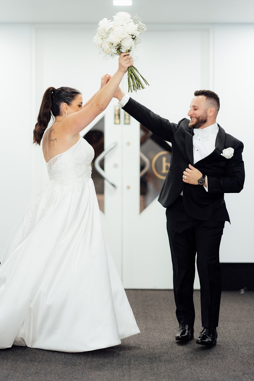 Bride in a white wedding dress and groom in a black tuxedo celebrating with a bouquet of flowers in an indoor setting at Donato reception centre in Adelaide