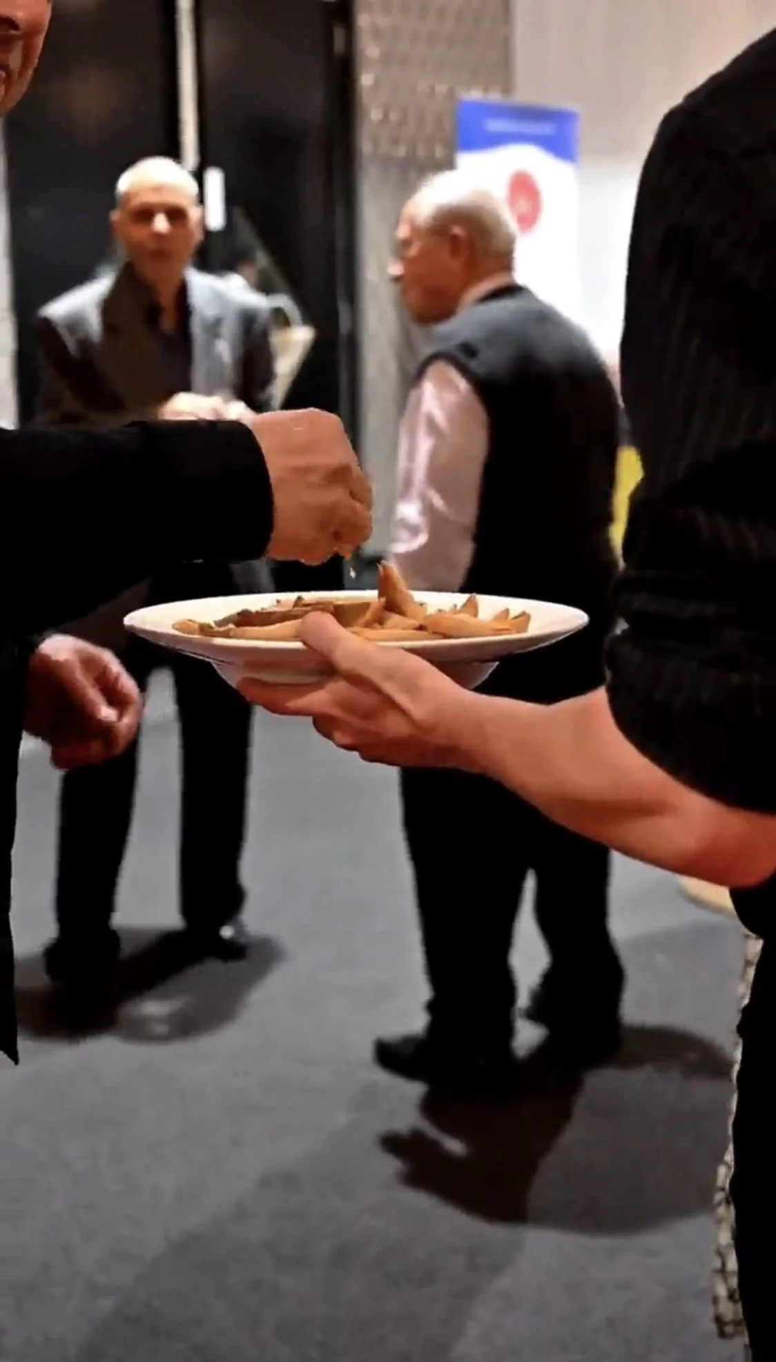Person holding a plate of food, possibly fries, at an event with people in the background.