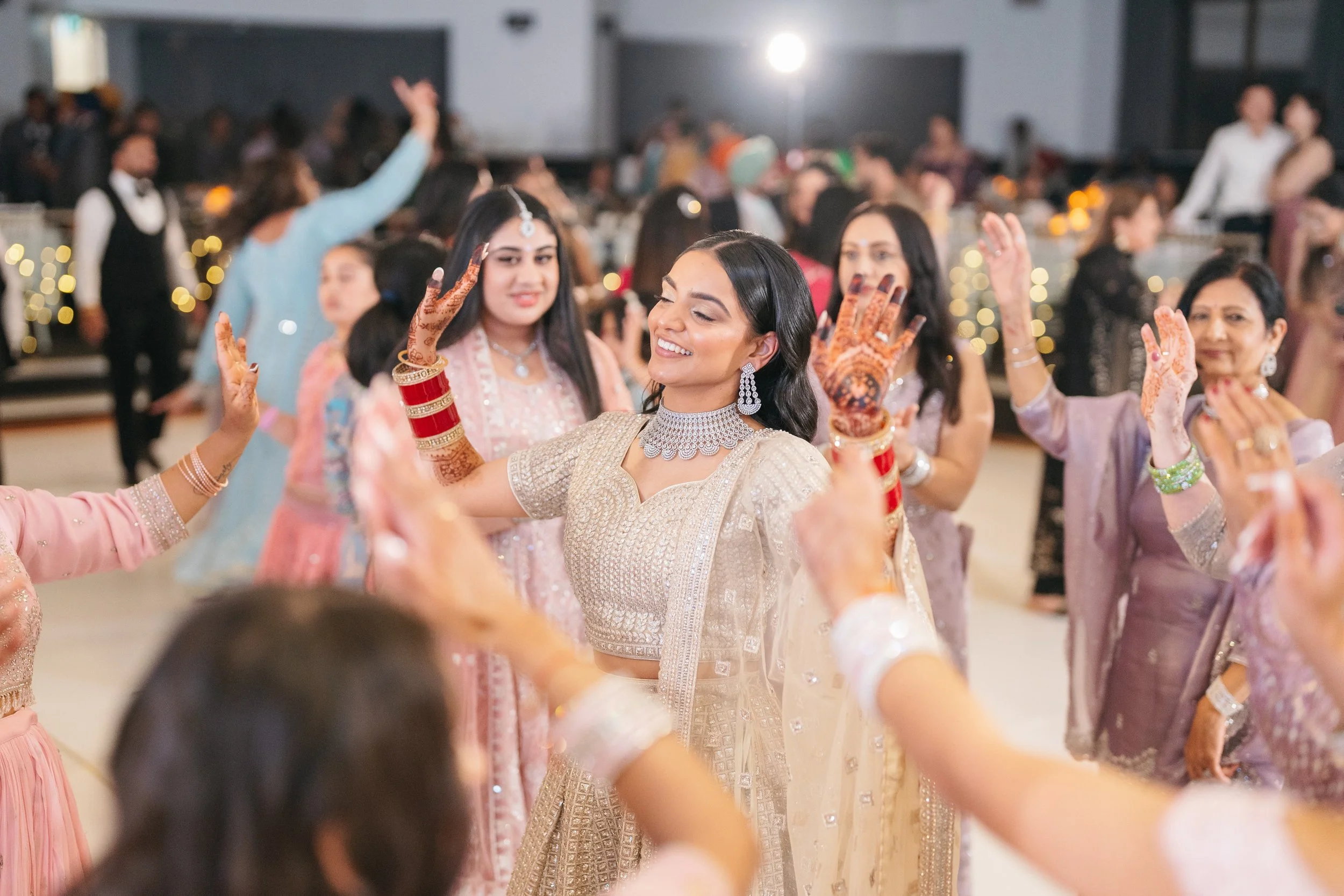 Women dancing and celebrating at a wedding reception, dressed in traditional Indian attire with henna on hands, jewelry, and smiles in a decorated venue.