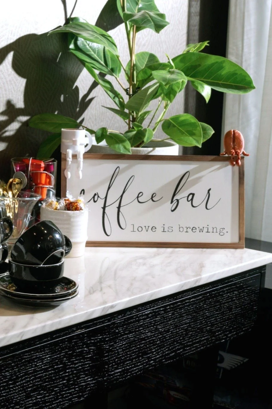 A coffee bar setup with a potted plant, black coffee cups, and a sign that says 'coffee bar, love is brewing', on a marble countertop near a window.