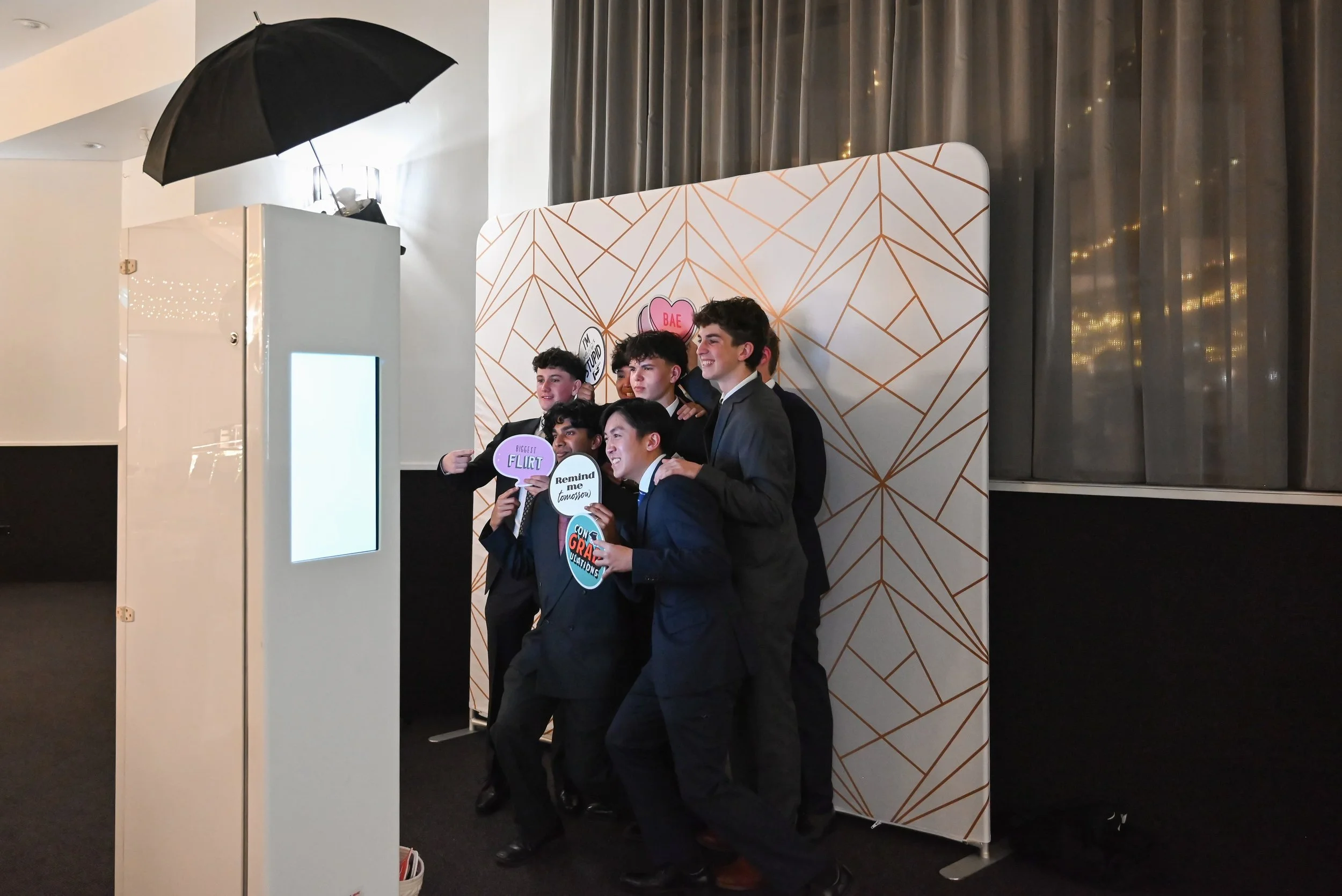 A group of young men dressed in suits taking a photo together at a photo booth with a geometric patterned backdrop, holding various signs.