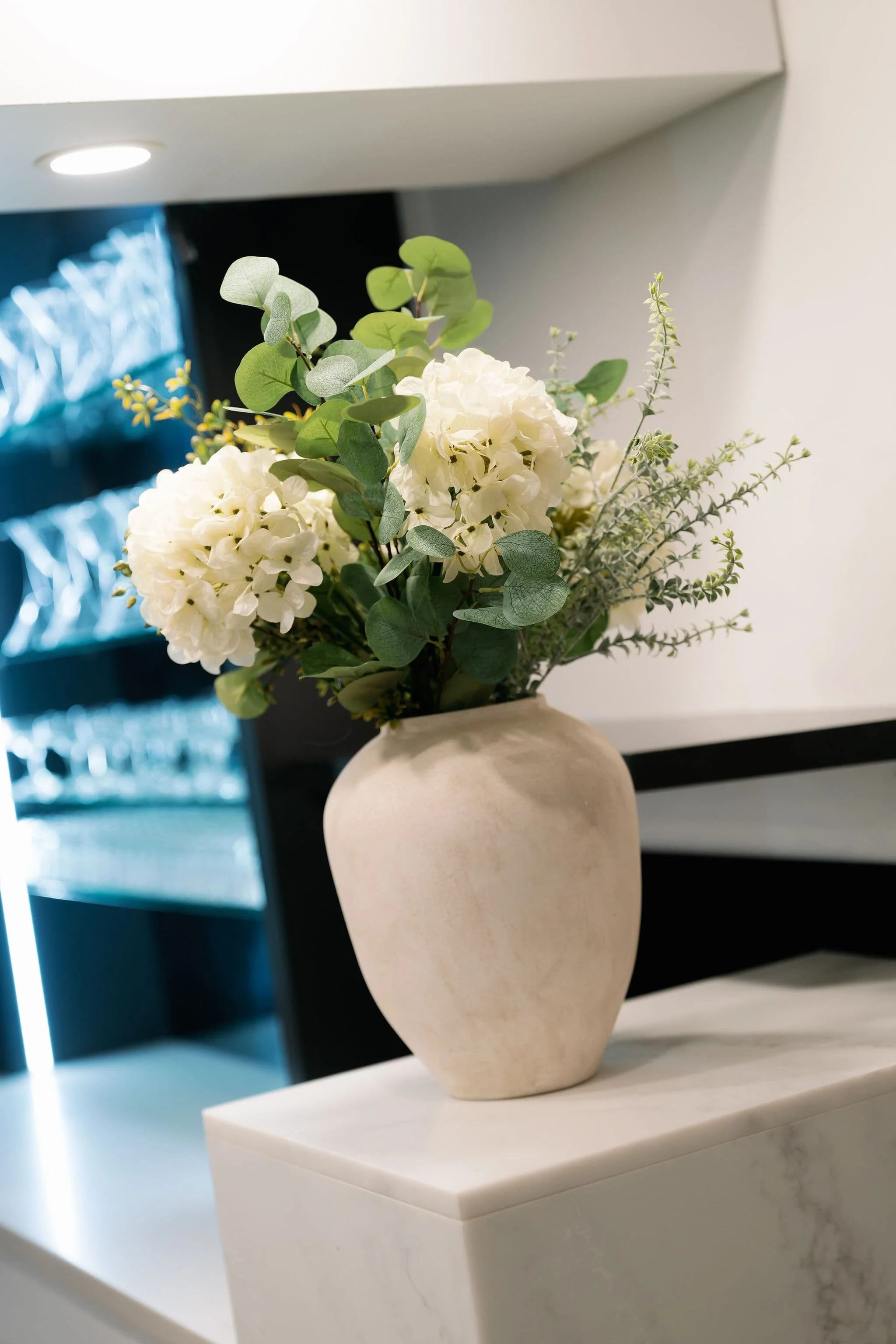A white ceramic vase with a bouquet of white hydrangeas and green foliage, placed on a white marble surface, in a modern interior setting.
